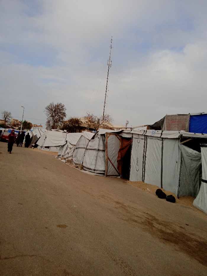 A row of makeshift tents lines a paved road, likely in a refugee camp setting. The tents are constructed from white and gray materials, some appearing more sturdy than others. A tall metal tower rises above the tents, possibly a communication or observation structure. Several people walk along the road past the tents. The sky is overcast. Overall, the image portrays a scene of displacement and temporary shelter.