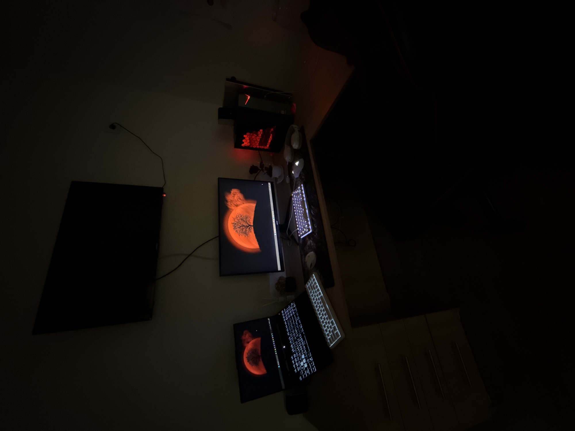 The image depicts a dimly lit home office setup. The focal point is a wooden desk with two monitors, a laptop, two keyboards, and two mice. The main monitor displays a vibrant orange tree against a similar-colored sunset or moon. The laptop, positioned to the left, also shows a similar red moon wallpaper. Both keyboards are illuminated with white backlighting. To the right of the main monitor, a black computer tower with red internal lighting is visible. A small potted plant and headphones are also present on the desk. Above the desk, a large black television is mounted on the wall. The room is predominantly dark, with the primary illumination coming from the screens and the computer tower's LEDs, suggesting it is nighttime or the room's lights are off. There is no visible indication of the specific location being in Krushev Dol, Bulgaria.