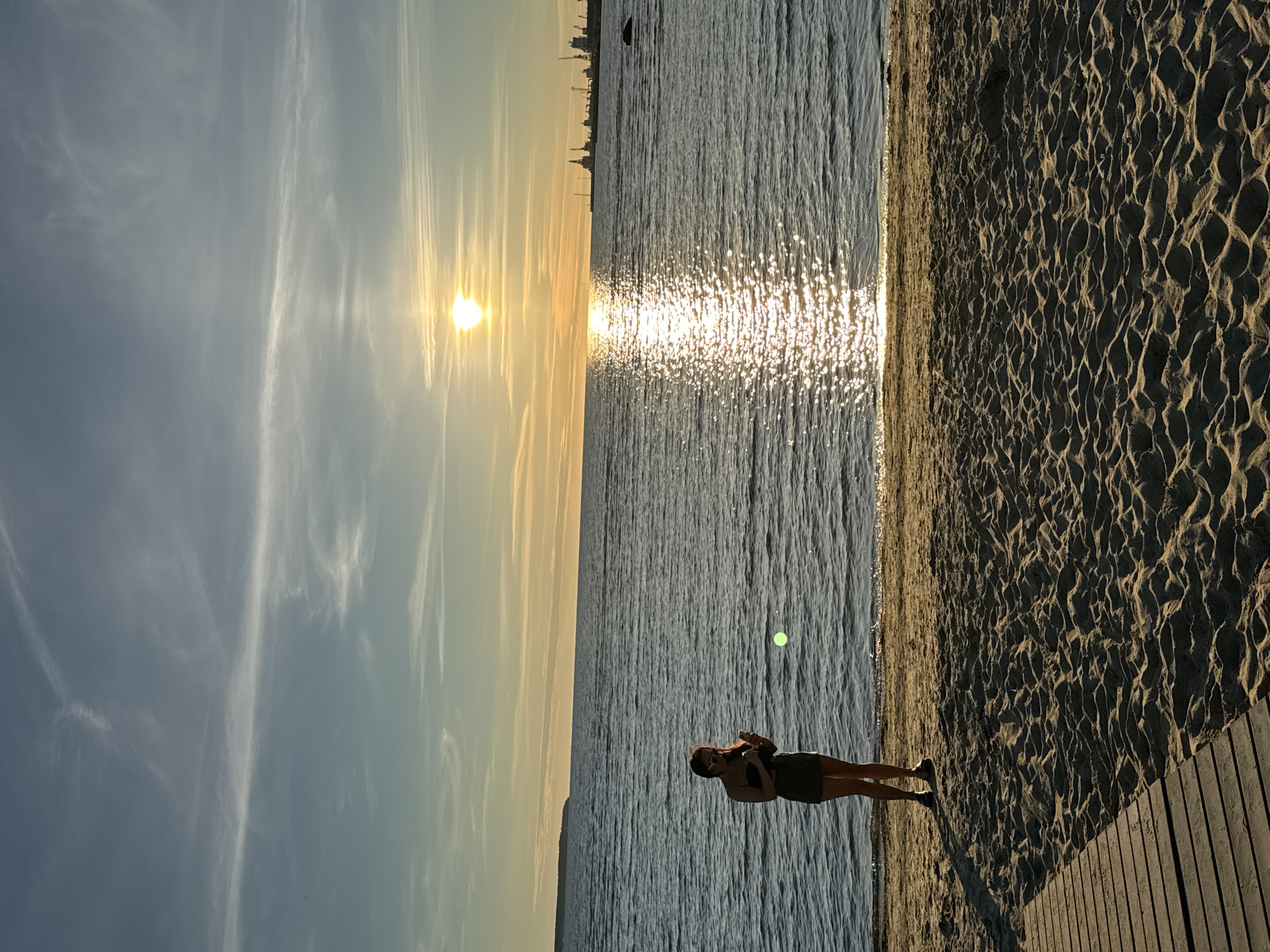 The image depicts a serene sunset scene at a beach in Tallinn, Estonia. A lone individual, a woman wearing a dark top and shorts, stands on the sandy shore, looking down at her phone. Her shadow stretches out on the sand, illuminated by the warm, golden light of the setting sun. The sun, a bright orb in the sky, casts a shimmering reflection on the calm waters of the Baltic Sea. In the distance, a silhouette of a distant landmass and industrial cranes on the horizon suggest the proximity of the city's port. The sky is adorned with wispy clouds, tinged with the colors of the sunset. A wooden boardwalk is partially visible in the foreground, leading onto the sandy beach. The overall atmosphere is peaceful and tranquil.