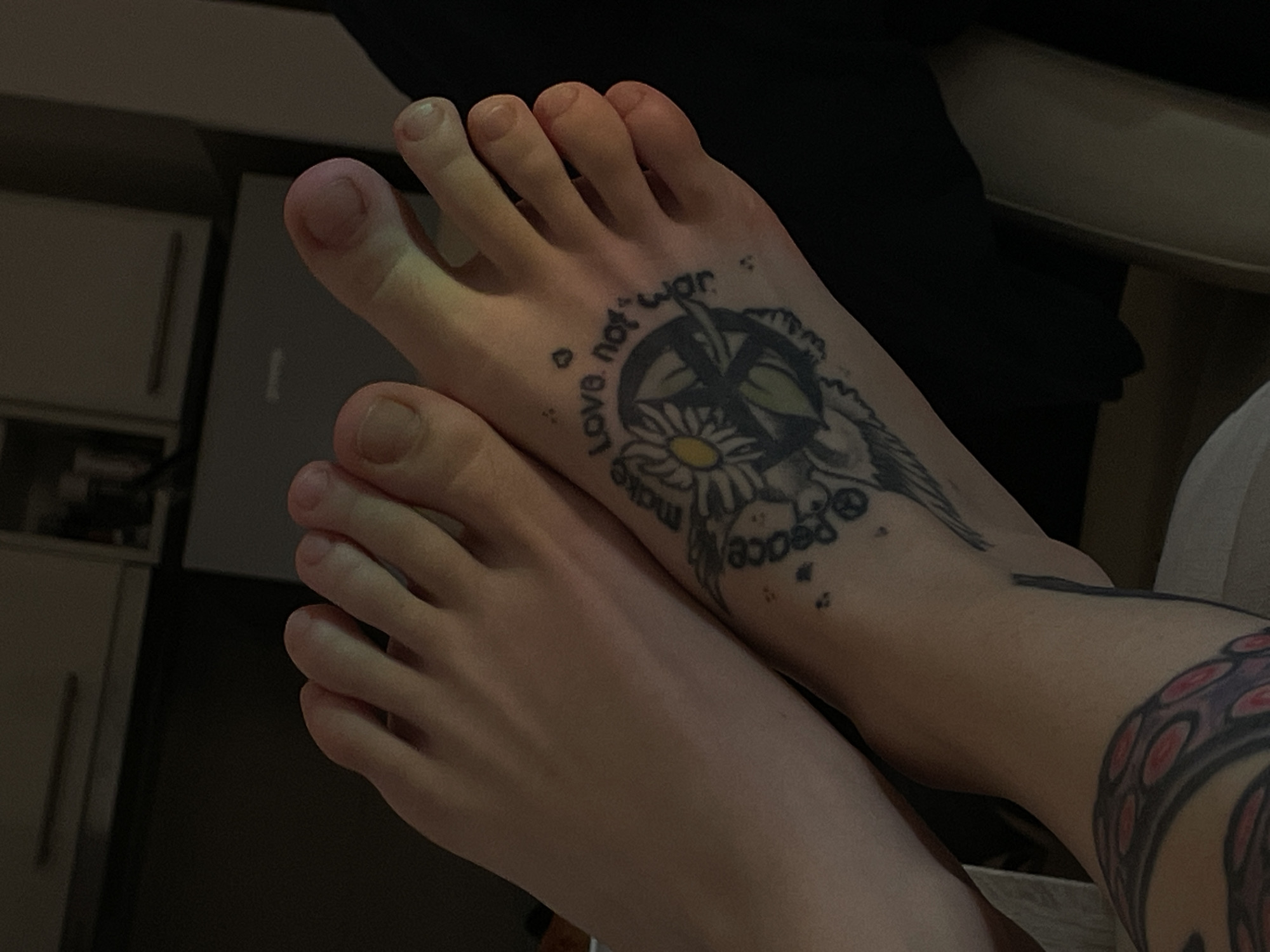 The image displays two bare human feet in a dimly lit indoor setting. One foot, positioned on the left, appears to rest slightly above or alongside the other foot, which is on the right. The foot on the right features multiple tattoos. A prominent tattoo on its dorsal surface is circular, depicting a daisy flower at its center, surrounded by a peace symbol, with the text "make love not war" inscribed around it. Adjacent to this, small, scattered dot or star-like tattoos are visible. Further down on the right ankle/lower leg, a larger tattoo is partially visible, depicting what appears to be an octopus tentacle with red and purple colored suckers. The background is dark and out of focus, revealing indistinct shapes in the upper left that resemble cabinets or appliances with horizontal handles. The immediate surface beneath the feet is light-colored and indistinct. There is no visual information within the image to determine the location in Zvezdara Urban Municipality, Serbia.
