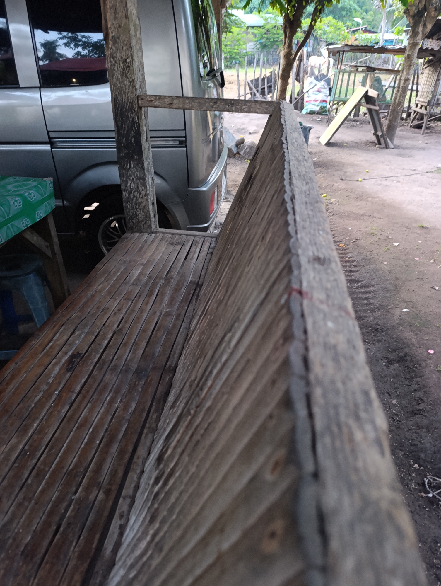 The image shows an outdoor scene, likely in a rural or semi-rural area, with a focus on a wooden bench constructed from thin slats. To the left, a portion of a silver van is visible. The bench is weathered and appears to be part of a simple outdoor seating area, possibly associated with a dwelling or a small establishment. In the background, there are trees, a wooden fence, and some makeshift structures, including what appears to be a small open-air shelter with some items underneath. The ground is dirt and appears somewhat uneven. The overall impression is one of a simple, utilitarian outdoor space.