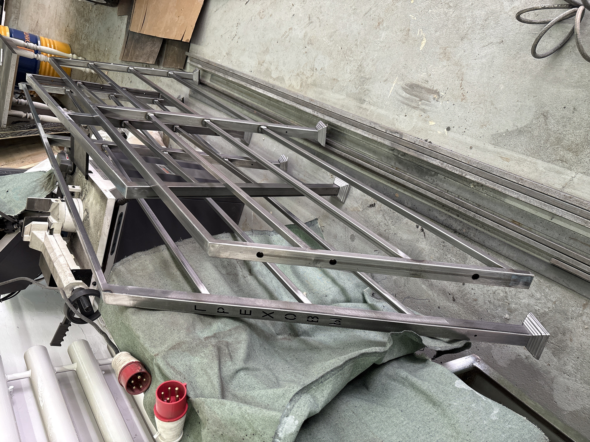 This media file captures an indoor industrial or workshop setting, likely in Chaikovsky, Russia. The central subject is a large, partially assembled metal framework, constructed from numerous rectangular steel or aluminum bars. This framework features a grid-like pattern with both horizontal and diagonal elements, and some components include pre-drilled holes and angular end caps or decorative sections. The metal appears newly fabricated, unpainted, and shows welding or joining points, indicating it is either in production or awaiting further assembly.
The framework rests partly on a large, light green or grey-green fabric covering an unseen surface, and partly on various pieces of workshop equipment. In the background, stacks of raw metal bars, similar in profile to those used in the main structure, are visible, suggesting an active fabrication environment. The floor is made of concrete, consistent with an industrial workspace, and displays some marks and debris.
To the lower left, two red and white industrial electrical plugs, each with multiple pins, are clearly visible, along with some white-painted metal pipes or bars. A black electrical cord is also present. A blue and yellow barrel, partially visible in the upper left background, carries some indistinct Cyrillic text, possibly "...СМИТН".
A prominent detail is the Cyrillic word "ЛАРЕХОВ" (LAREKHOV) stamped or etched into one of the horizontal bars of the metal framework. This text likely serves as a brand name, project identifier, or manufacturer's mark. The scene depicts a phase of metal construction or fabrication, with raw materials and components in various stages of completion. No individuals are present in the image, and the time of day is not discernible from the indoor lighting.