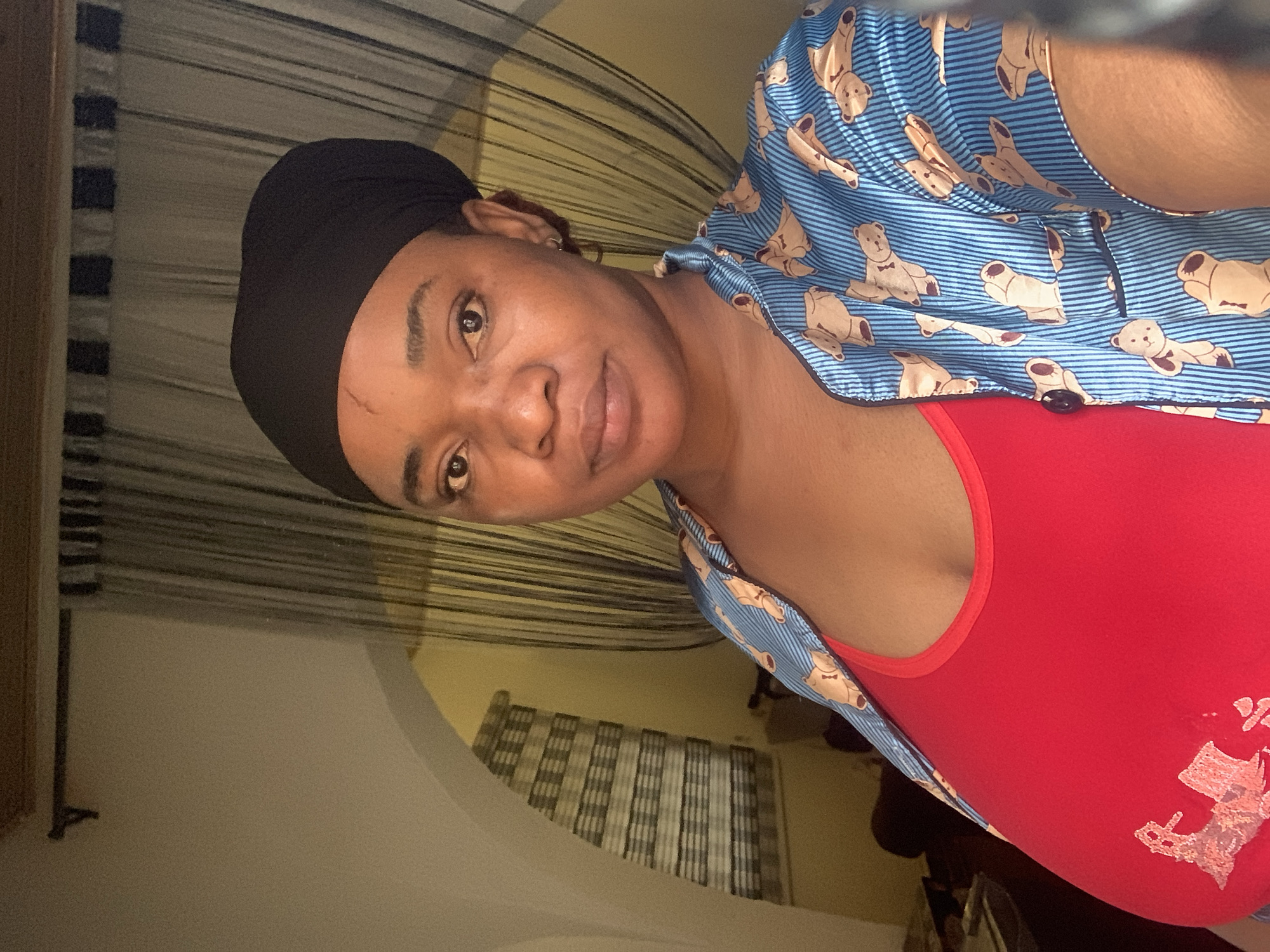 A woman is captured in a close-up portrait or selfie, rotated to a tilted orientation. She has a warm skin tone and wears a black headwrap or cap. Her brown eyes look directly at the viewer, and she has a gentle, soft smile. A notable scar is visible on her forehead, positioned above her left eyebrow.
She is dressed in a blue and white striped short-sleeved button-up shirt featuring a pattern of light brown teddy bears. Underneath, she wears a bright red top. A faded or peeling graphic is present on the lower right side of the red top, but its details are indistinguishable.
The background suggests an indoor setting, likely a room. Behind her, there are horizontally striped curtains or blinds in shades of dark and light, possibly black and white or grey. Another window with a similar striped blind pattern is visible further back in the room. The lighting is soft and ambient, characteristic of indoor lighting conditions. The overall impression is casual and personal. The context provided indicates this image was taken in Barakin Akawo, Nigeria.