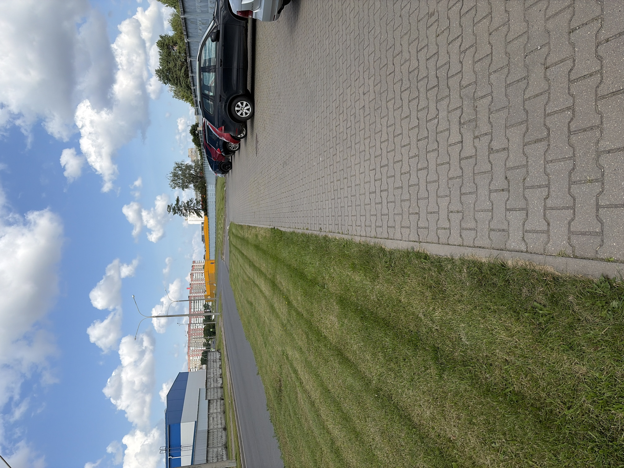 The image captures a view of a parking lot and a roadside area in Navadworski Selski Soviet, Belarus. In the foreground, a paved walkway made of interlocking gray bricks runs parallel to a grassy verge. The grass has distinct mowing patterns, with lighter green stripes indicating recently cut areas. Beyond the grass is an asphalt road.
On the right side of the road, several cars are parked in a line. The closest car visible is a black SUV, followed by a red car, and then other vehicles partially obscured. Beyond the parked cars, a concrete fence runs along the property line, with trees and foliage visible behind it.
In the background, a row of multi-story residential buildings with red brick facades can be seen. To the left, a large building with a blue and white exterior is partially visible. The sky is a bright blue with scattered white, fluffy clouds. Two streetlights are visible along the roadside, one taller than the other. The overall scene depicts an urban or suburban setting on a clear day.