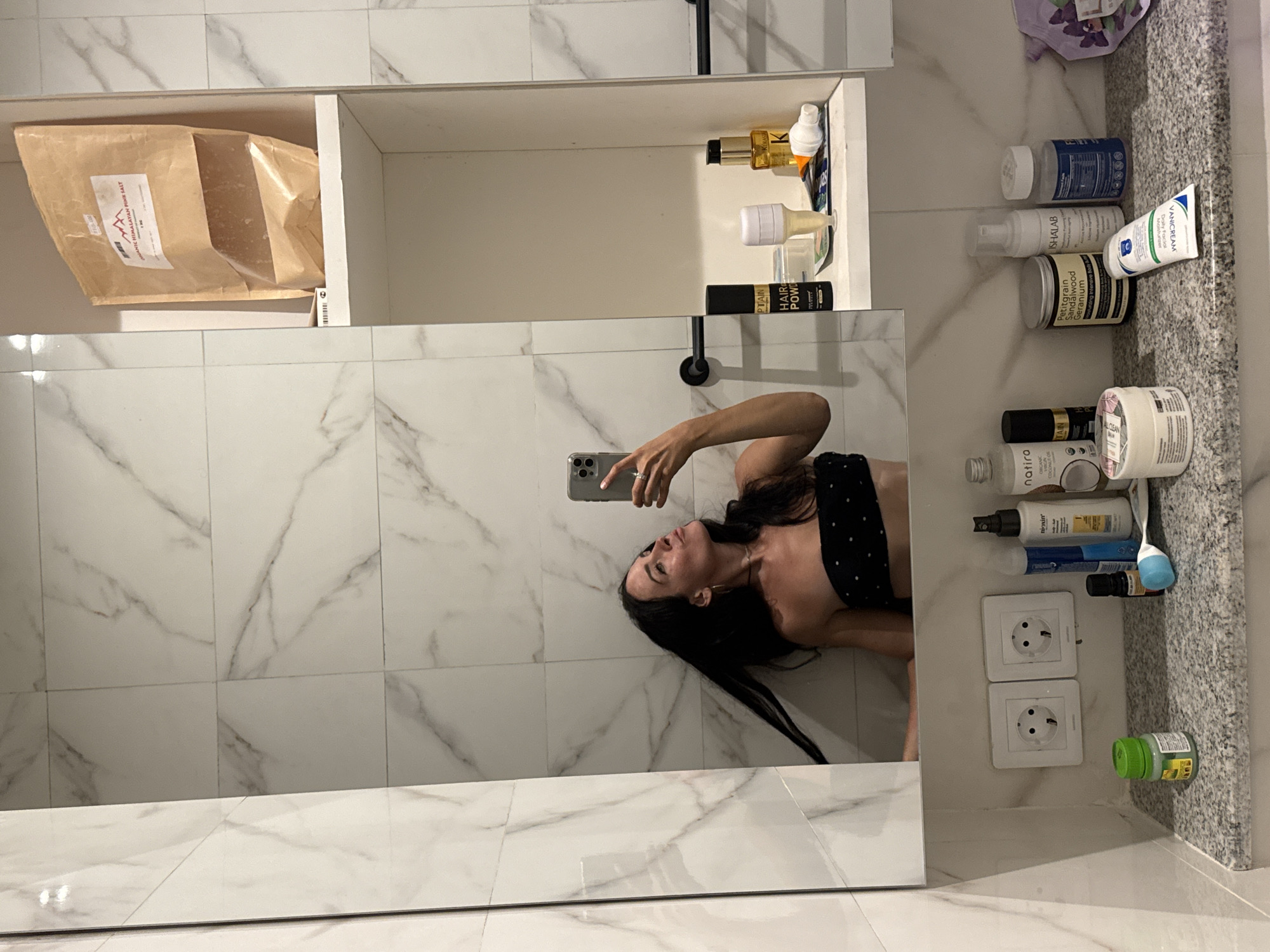 A woman is taking a mirror selfie in a bathroom. She is wearing a black bandeau top with white polka dots. She has long dark hair that is being swiped to the left, and she is looking up and to the right, puckering her lips. Her right arm is raised, holding a silver smartphone.
The bathroom features white marble-patterned tiles on the walls and a white countertop. Above the countertop, there is a large mirror that reflects the scene. To the right of the mirror, a white shelf is visible, containing several items. On the top shelf is a large brown paper bag with a label that reads "ORGANIC HIMALAYAN PINK SALT." Below that, on the shelf, are a black spray bottle labeled "CAPTAIN HAIR POWER," a small bottle with a white dispenser, and a golden bottle of liquid.
On the countertop below the mirror, there are several toiletries and bathroom products. These include two white electrical outlets, a small green bottle, a black bottle, a blue and white spray bottle, a white bottle with a pump dispenser, a cylindrical container with a yellow label, a white tube of cream, a white bottle with a blue label, and a small purple package with a floral design. The countertop appears to be made of speckled granite.