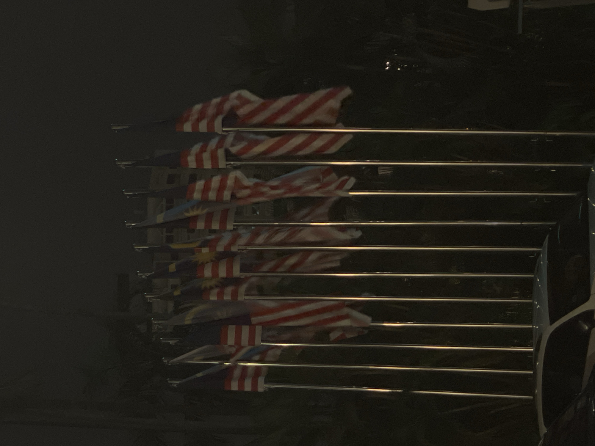 The image shows a row of Malaysian flags flying at night, illuminated by artificial lights. The flags are mounted on tall, silver poles, and some of them are fluttering in the wind. The background is dark and blurry, with some faint shapes that might be trees or buildings. In the foreground, the top of a white car with a sunroof is visible. The scene appears to be an outdoor public space, possibly in Kuala Lumpur, Malaysia, given the presence of Malaysian flags.