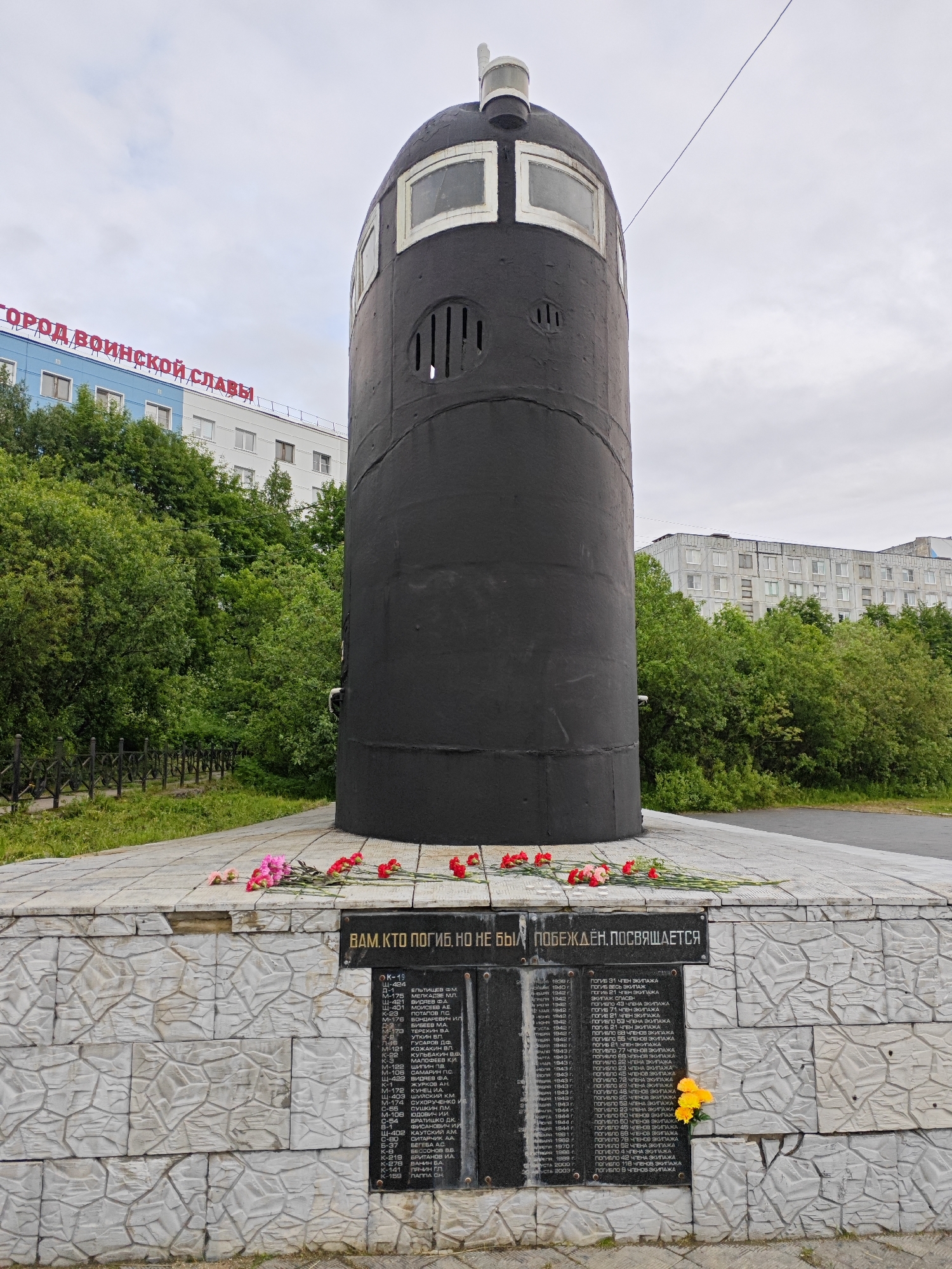 This is a photograph of a memorial in Polyarny, Russia, dedicated to those who perished but were not defeated. The memorial features a large, dark cylindrical structure resembling a submarine conning tower, with windows at the top. Below this structure is a stone base with black plaques listing names and dates, likely of fallen soldiers or sailors. Red and pink flowers are placed on the base and a yellow bouquet is nestled in a crevice of the stone. In the background, trees and multi-story buildings are visible, including one with the red lettering "ГОРОД ВОИНСКОЙ СЛАВЫ" (City of Military Glory). The sky is overcast, suggesting a cloudy day. The overall mood is somber and respectful, commemorating a significant historical event or loss.