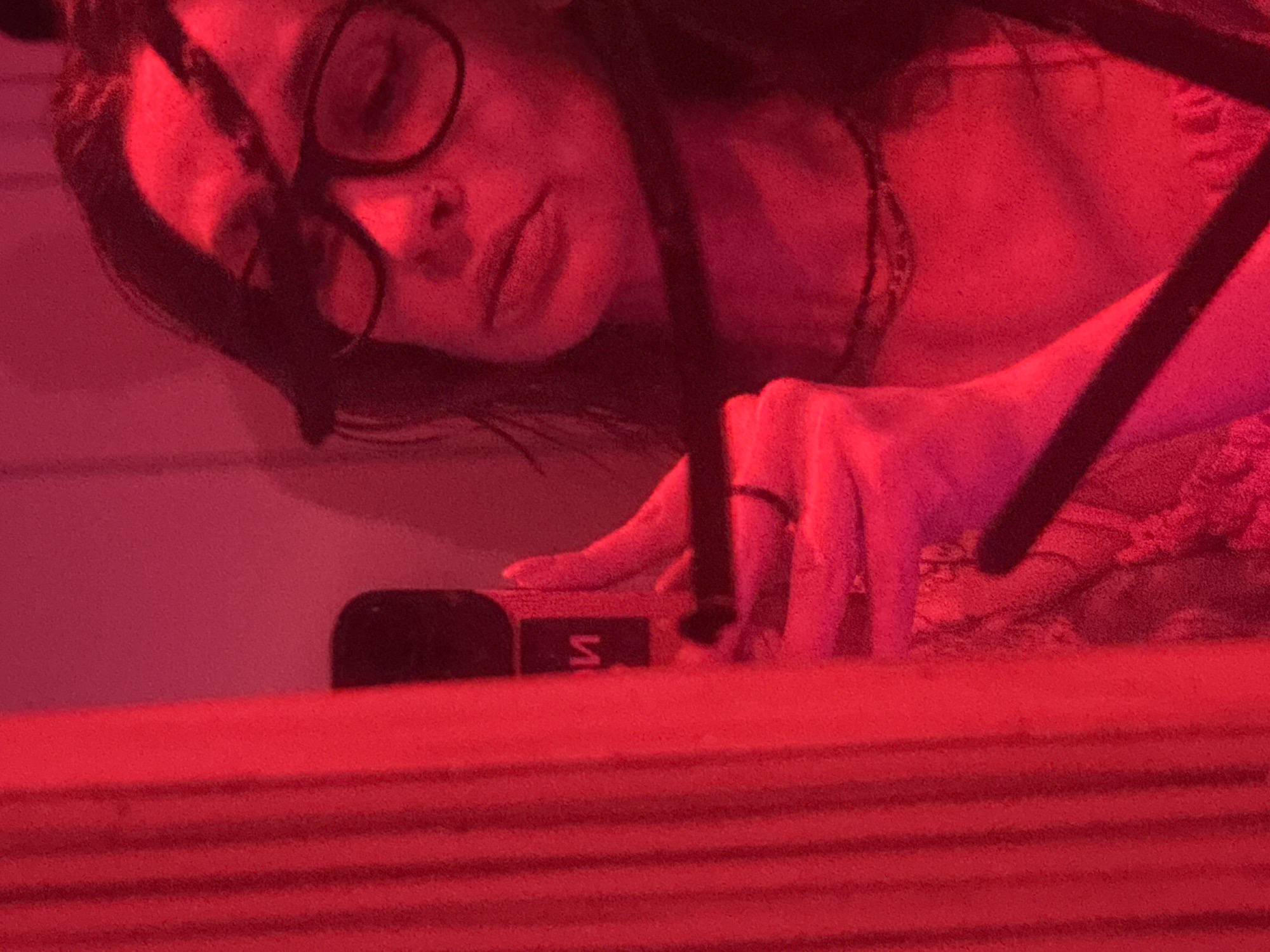 The image captures a person indoors, bathed entirely in strong red lighting. The primary subject is a person with dark hair and glasses, positioned in the upper left quadrant of the frame, looking downwards or towards an object. Their upper body, including their face, neck with a necklace, and right hand, is visible.
In the center-left, the person's right hand rests near a small, black, rectangular object. This object features a distinct white "S" logo on its side. A ring is visible on the person's right ring finger. In the foreground, a textured surface with horizontal lines, possibly a table or desk, occupies the bottom half of the image. Dark, angled bars, likely part of furniture or a structure, are present in the upper right.
The pervasive red light makes it difficult to ascertain true colors or time of day, but suggests an artificial, potentially evening or night-time setting, creating a moody or intimate atmosphere. The person's expression appears calm or contemplative. There are no direct visual cues within the image to confirm the location as Ramat Gan, Occupied Palestine, as provided by the context.
