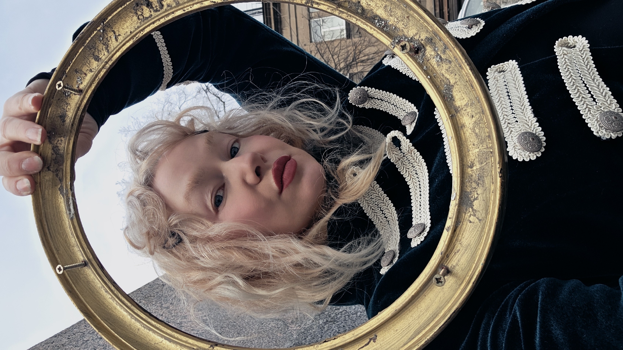 A low-angle shot shows a woman holding a gold, circular frame, framing her face. The woman has blonde, curly hair and wears a dark-green velvet jacket adorned with white braid and silver buttons. Her makeup includes dark lipstick and blush. The frame is old, with visible wear and screws. The background shows a building with windows and bare trees. Based on architectural style and the sky's hue, the location is likely in the city of Saint Petersburg, Russia.