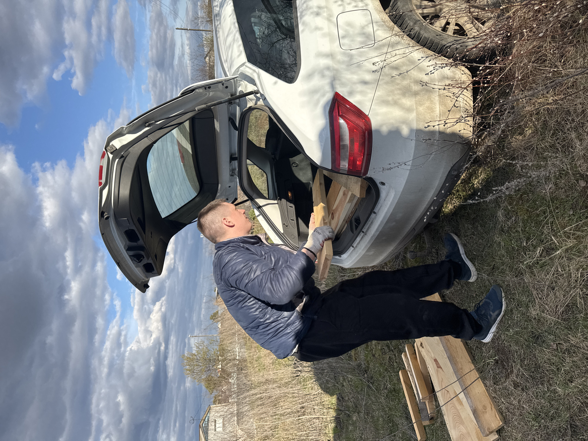 A man is engaged in loading wooden planks into the open trunk of a white hatchback car, set in what appears to be a rural or semi-rural environment in Noviy, Russia. The image is oriented sideways, captured during daytime with bright sunlight.
The man has fair skin and short, light-colored hair. He is dressed in a dark blue puffer jacket, dark trousers, and dark sneakers with white soles. He wears light-colored work gloves, with the text "EKF" faintly visible on his right glove. He holds a piece of unfinished wooden plank, carefully placing it into the trunk, which already contains several other similar pieces of lumber.
The white car shows signs of dirt or dust, particularly around the rear wheel and lower body, suggesting travel on unpaved roads. It is parked on dry, sparse grass mixed with bare bushes, some of which exhibit early spring pussy willows. The sky above is partly cloudy, featuring both bright blue patches and white to grey clouds. In the background, distant structures and utility poles are visible, contributing to the rural atmosphere. The overall scene captures a practical activity under clear weather conditions.