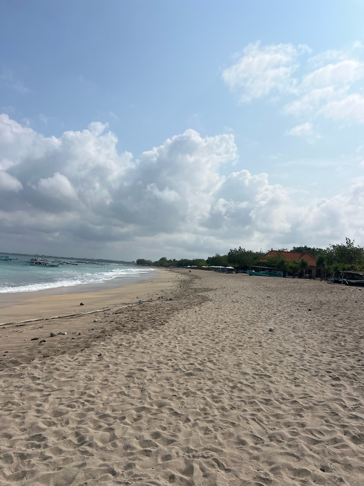 The image depicts a sandy beach in Kuta, Indonesia, under a partly cloudy sky. The foreground is dominated by textured sand, with a thick rope running horizontally across the scene, partially buried. Further back, the shoreline meets the ocean, with gentle waves breaking on the sand. Several traditional Indonesian fishing boats, known as perahu, are visible anchored in the turquoise water. Along the coastline, trees and buildings are scattered, including structures with orange-tiled roofs. The scene suggests a typical day at a beach resort area, with a mix of natural elements and human activity.