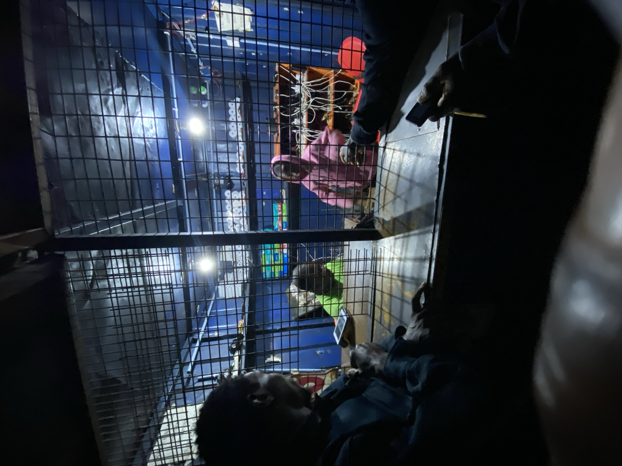 The image depicts a scene inside a caged enclosure, likely a small business or service booth, in Bukuru, Nigeria. Two individuals are visible within the enclosure. On the left, a person wearing a bright green t-shirt is partially visible, possibly interacting with something on a counter. To their right, a person in a pink hooded sweatshirt is standing and looking towards the right side of the enclosure. Behind them, shelves are stocked with various items, and electrical wiring is prominently displayed. A bright light illuminates the back of the enclosure.
Outside the enclosure, several people are visible from the chest down, suggesting they are standing at a counter or window. One person on the left is wearing a dark jacket and has their hand resting on the counter. Another person on the right is holding a mobile phone. The enclosure itself is constructed with metal bars, creating a secure or partitioned space. The overall lighting is dim, with artificial lights providing illumination within the enclosure. The environment appears to be an urban setting, given the mention of Bukuru, Nigeria.