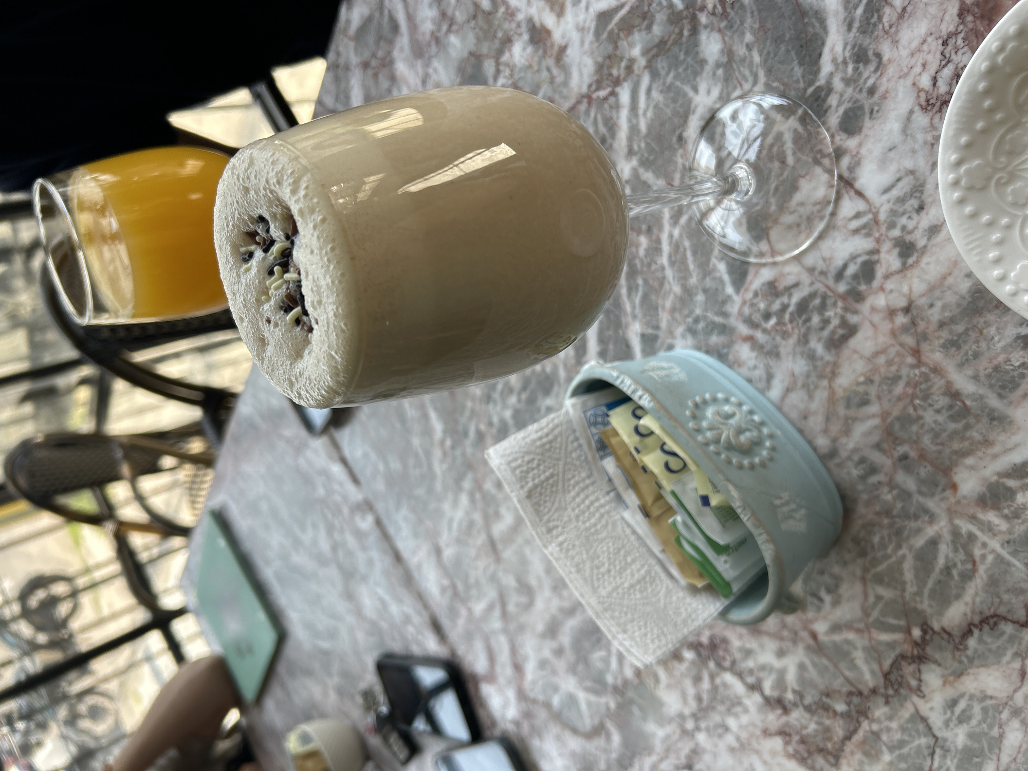 This image presents a top-down view of a marble-patterned table set with drinks and accessories, implying a cafe or casual dining scenario during the daytime due to the bright lighting.
The main subject is a tall, stemmed glass, positioned horizontally in the frame, containing a creamy, light-colored beverage, possibly a milkshake or a frothy coffee drink. The top of the drink is adorned with a textured layer of small, dark and white sprinkles or seeds.
In the blurred background to the left, another glass holds a bright orange liquid, likely orange juice. To the right of the prominent drink, a light blue, rectangular ceramic holder with an embossed floral or scroll pattern contains several sugar packets of various colors (yellow, green, white) and a folded white paper napkin. Some text is visible on the sugar packets but is largely unreadable; fragments like "S..." and "co" are faintly discernible.
A portion of a white plate with a raised, decorative pattern is visible on the far right. The table itself is made of marbled stone, featuring a mix of grey, white, and reddish-brown veining. The background is out of focus, showing dark, possibly metallic, furniture or structural elements, but no people are visible in the scene. The overall impression is one of a relaxed refreshment setting.