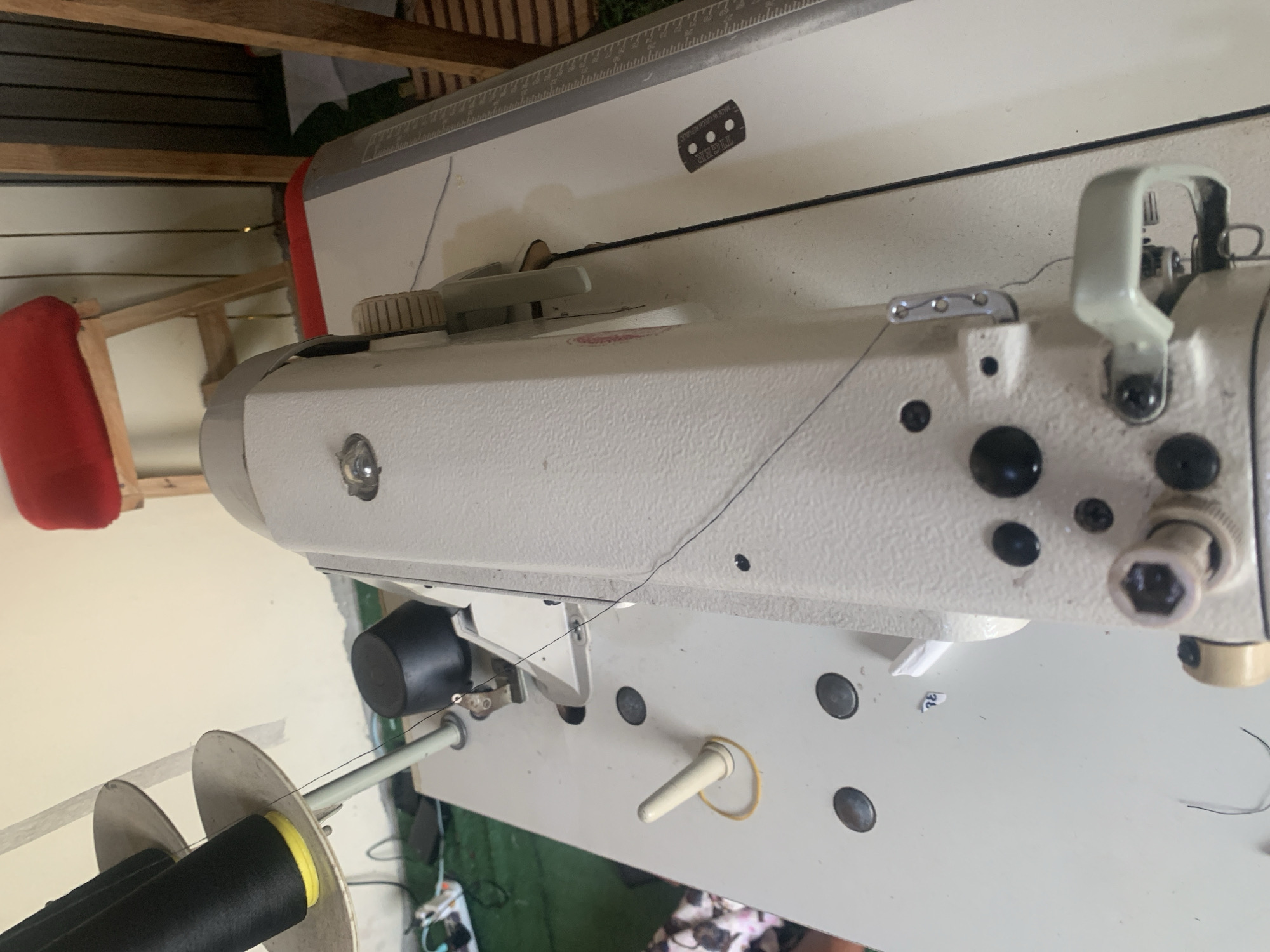 The image shows a close-up, high-angle view of a sewing machine on a white table. The sewing machine is cream-colored and appears to be industrial-grade, with various metal and plastic components visible, including a presser foot, handwheel, and thread guides. A black thread is visible, running from a spool on the left side of the frame, down towards the sewing machine. The spool itself is black with a yellow band near its base. In the background, a red-upholstered stool with wooden legs is partially visible, along with a portion of a wall. The setting is likely a workshop or a home sewing area. The image does not provide specific location details within Jauro Yinu, Nigeria, beyond what can be inferred from the presence of a sewing machine, suggesting a domestic or commercial context for textile work.
