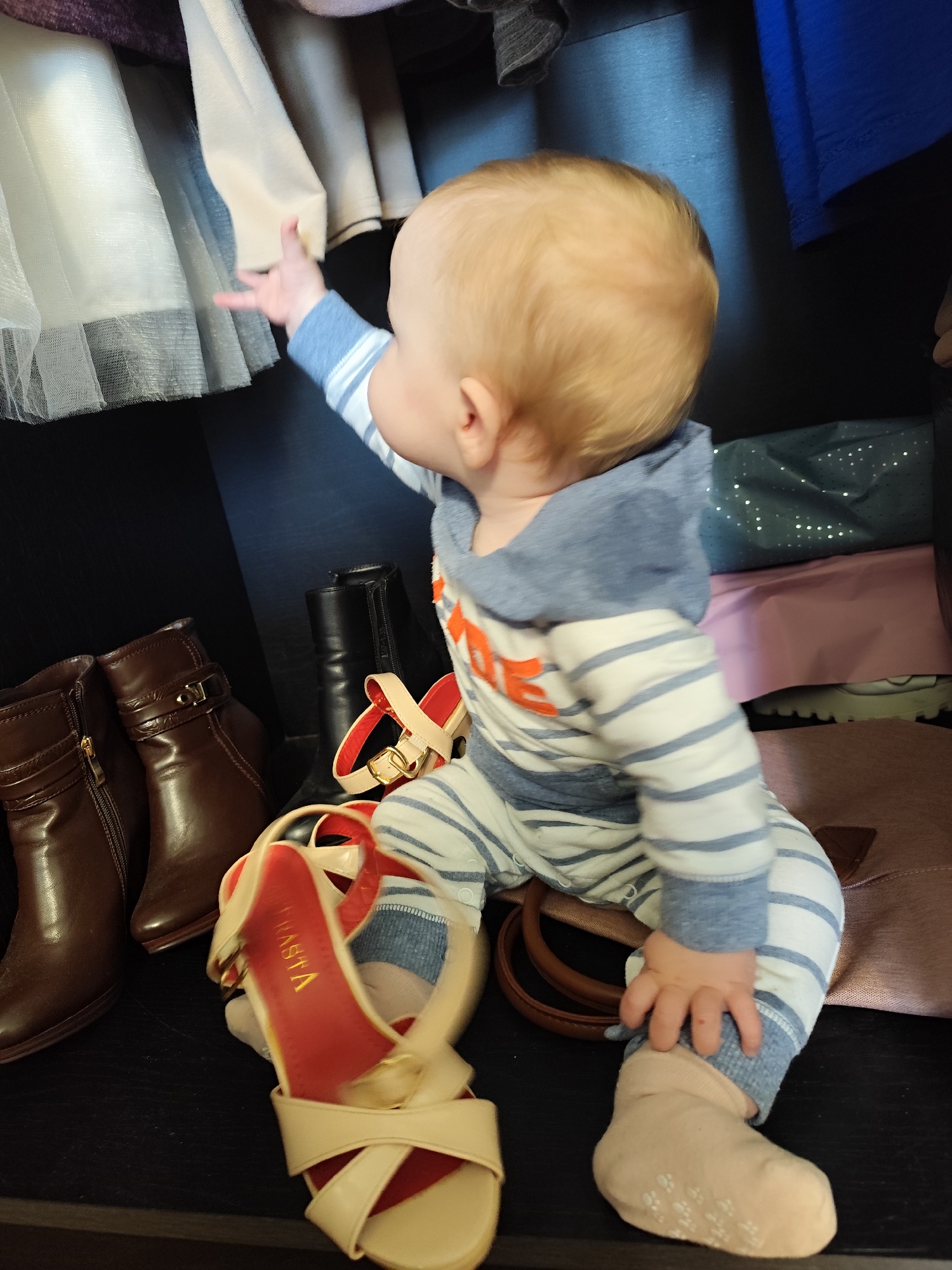 A baby sits in a closet surrounded by shoes and clothing. The baby wears a striped hooded jumpsuit and socks, and reaches up towards clothing hanging above. Brown and black ankle boots are visible, along with tan and red high-heeled sandals. Bags and wrapped gifts are behind the baby. The location is indoors, in a closet. There is no evidence to suggest that the location is in the city of Фоки, Russia.