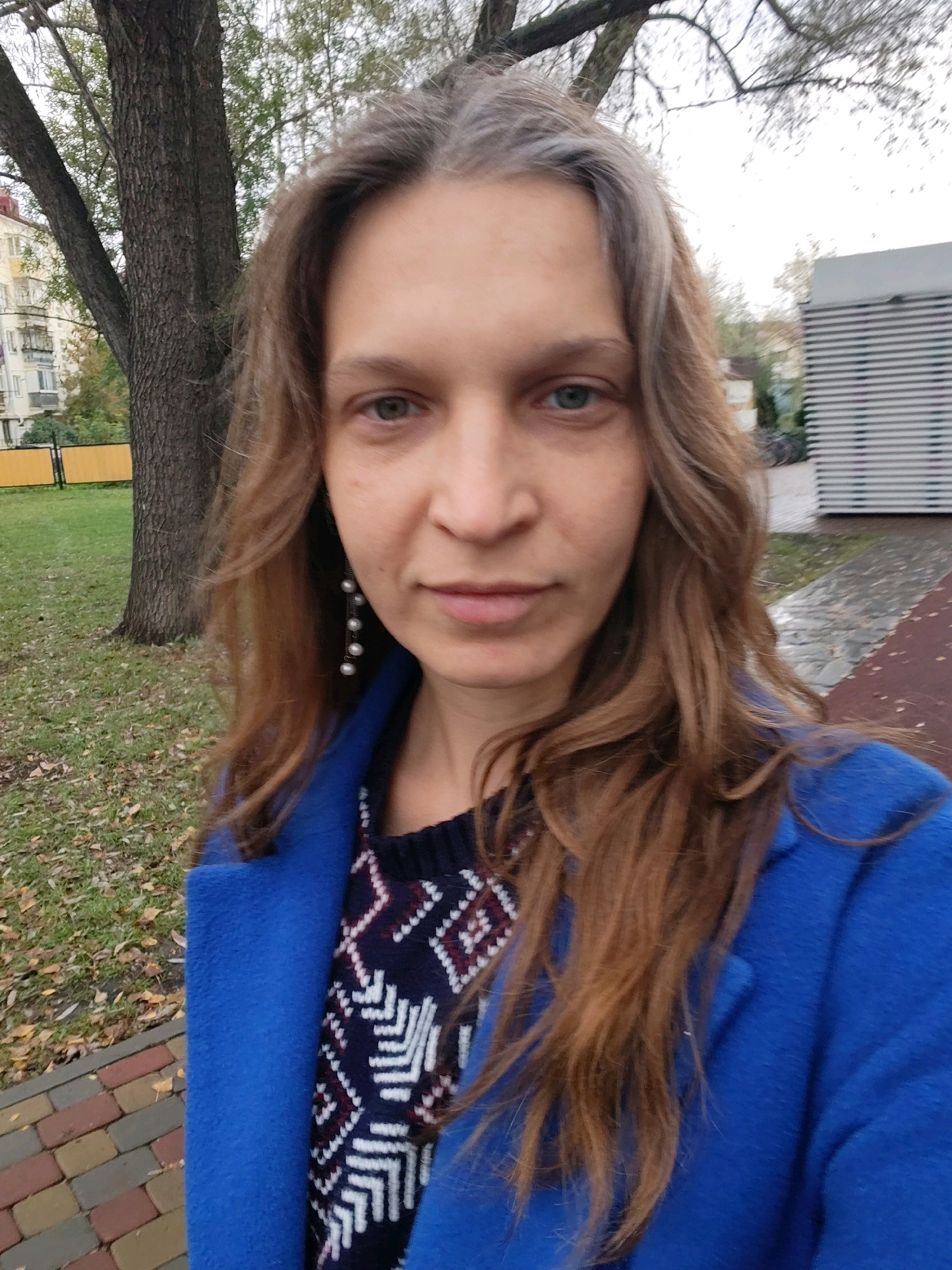 A woman with long, wavy brown hair and graying roots, wearing a blue coat over a patterned sweater and pearl earrings, is taking a selfie outdoors. She is standing in front of a tree with a textured trunk, a green lawn with fallen leaves, and a paved walkway with a reddish-brown border. In the background, there is a building with horizontal siding and a multi-story building with balconies. The scene appears to be in an urban or suburban park setting in the city of Kurgan, Russia, as indicated by the architecture and vegetation typical of the region.