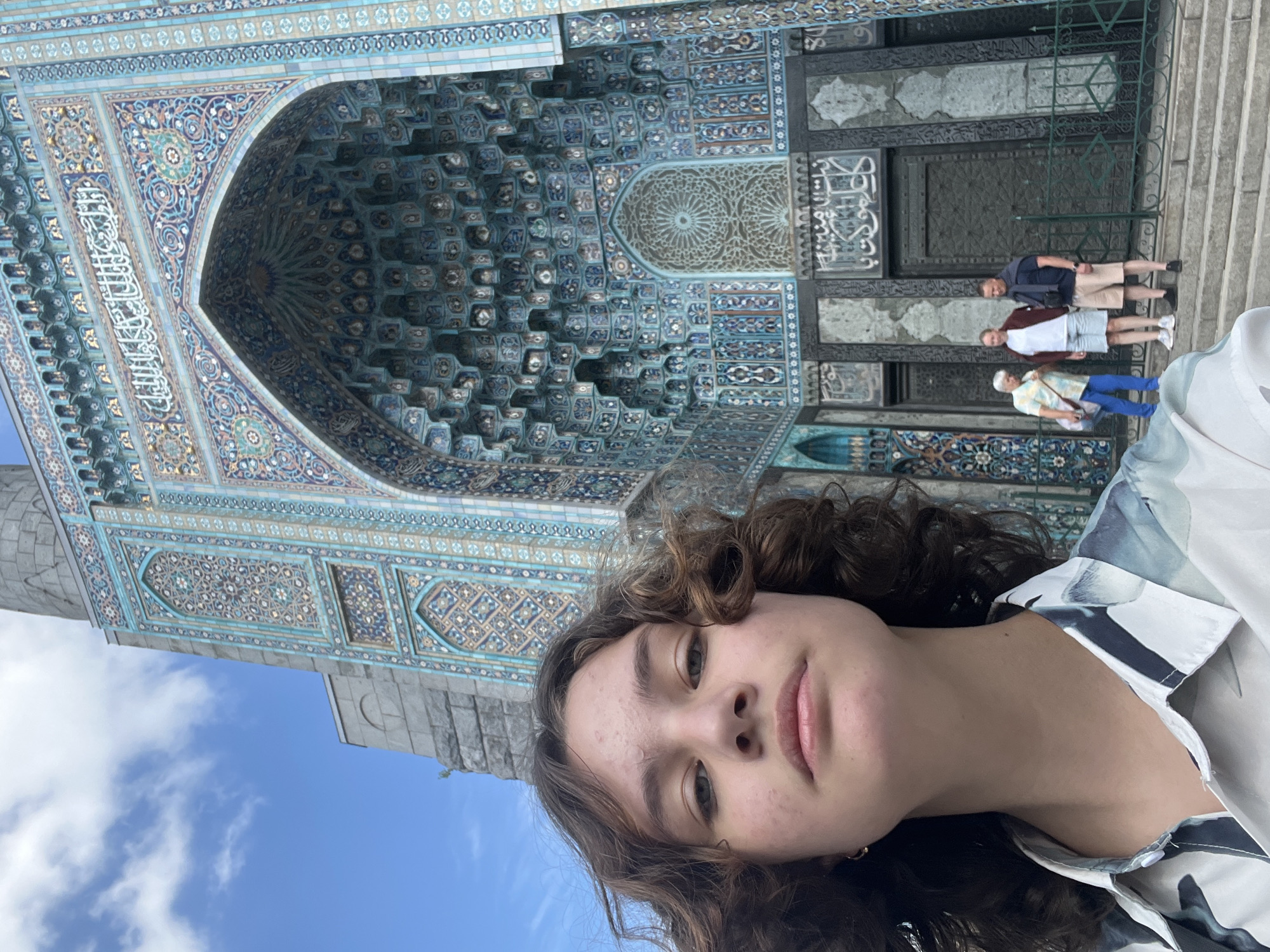 A young person with curly brown hair takes a selfie in front of a mosque in Saint Petersburg, Russia. The mosque has intricate blue and gold tilework. The person is wearing a light-colored shirt with a floral or abstract pattern. Behind the person, three other individuals are standing on a set of stone steps in front of the mosque's entrance. The sky is partly cloudy.