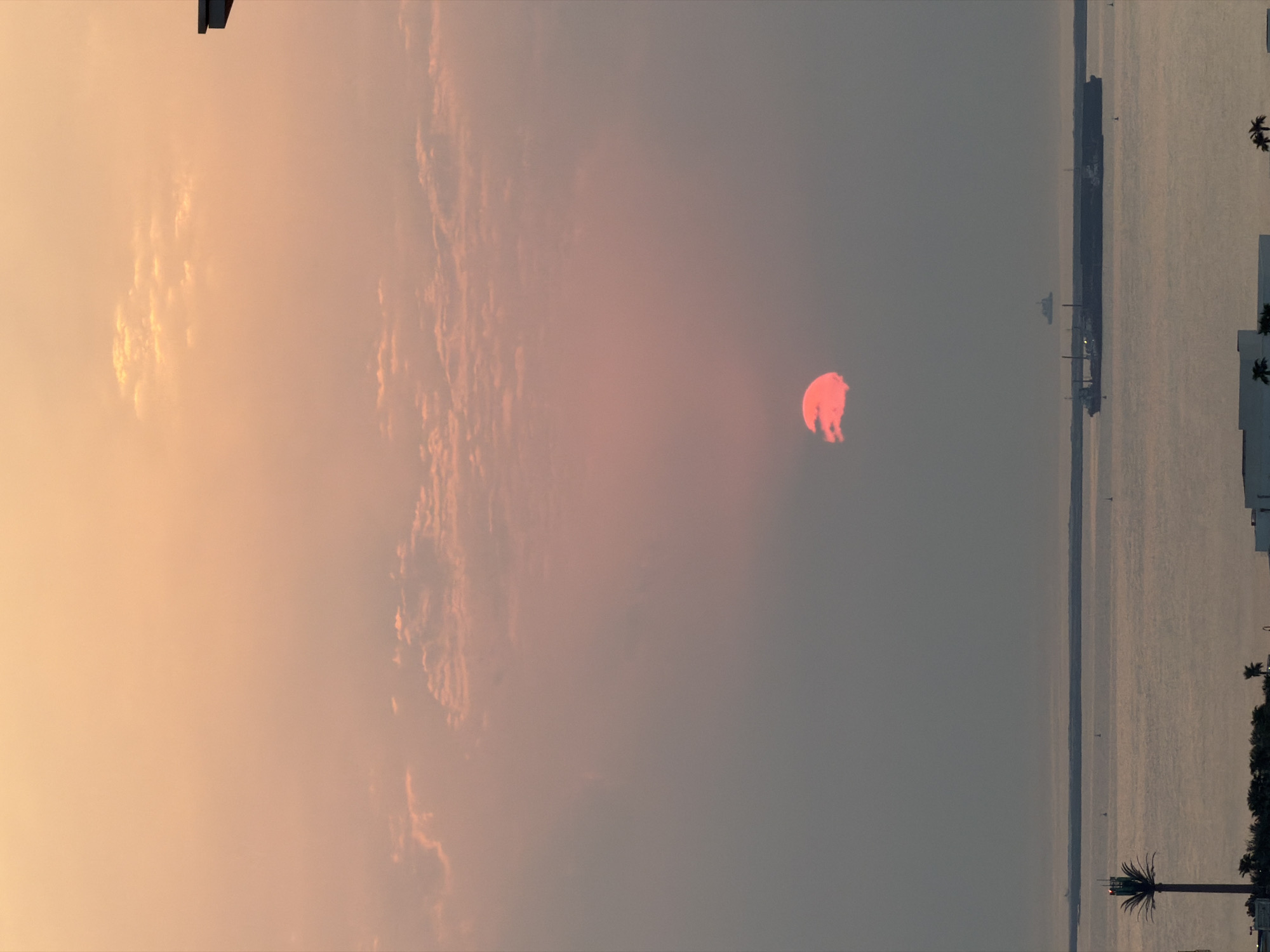 A sunset is captured over the water in Lusail, Qatar, casting a soft orange and pink glow across the sky. The sun, a vibrant red orb partially obscured by a cloud, hangs low on the horizon. The water below is calm, reflecting the muted colors of the sky, and a large cargo ship is visible sailing across it. In the foreground, silhouetted against the water, are buildings and palm trees, suggesting a coastal city setting. The overall atmosphere is serene and tranquil.