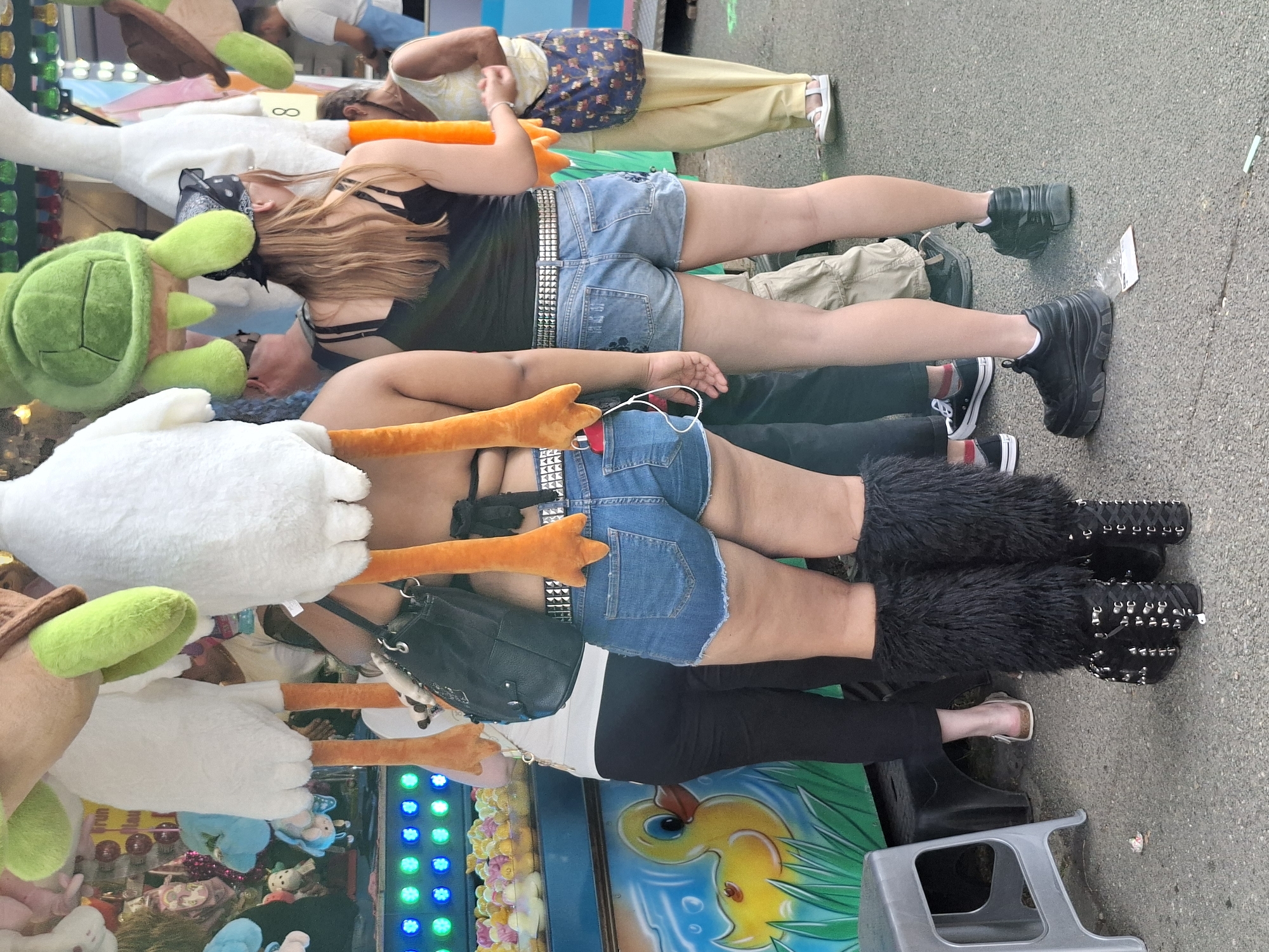 The image captures a scene at a street fair or carnival in Wiesbaden, Germany. In the foreground, a person wearing denim shorts and black furry leg warmers over studded boots stands with their back to the camera. They are holding onto a plush duck toy with orange feet and a white body. To their left, another person is visible from the chest down, wearing black pants and a white shirt, also holding a plush toy.
Behind them, a stall with various plush toys is set up, including a green turtle, and other duck-like figures with orange feet. The stall is decorated with colorful lights. In the background, other people are milling about, including a woman wearing a yellow top and a patterned bag, and a man in a white t-shirt and blue pants. The ground appears to be paved. The overall atmosphere suggests a lively, outdoor event.
