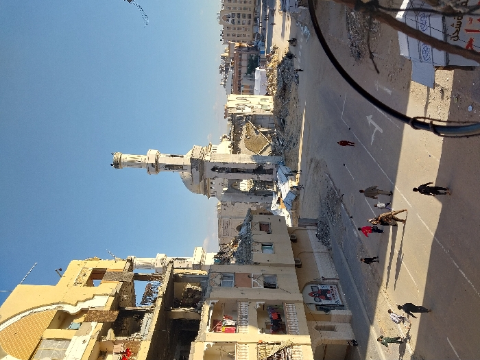 The image depicts an urban street scene in Khan Yunis, Palestinian Territory, under a clear blue sky. In the background, a large mosque is severely damaged, with its dome partially collapsed and extensive structural damage to its main body, though its minaret remains largely upright with damage visible at its top. Piles of rubble surround the mosque. To the left, a multi-story building exhibits heavy damage, with upper floors exposed, walls missing, and structural elements visible; some balconies still have laundry hanging, and a mural depicting red and black figures is painted on a lower wall. The foreground features an asphalt street with white painted road markings, including an arrow. Approximately 15 to 20 individuals, a mix of adults and children, are present on the street, walking or standing. Their shadows are distinctly cast, indicating bright daylight. Further damaged buildings and scattered debris are visible towards the background.