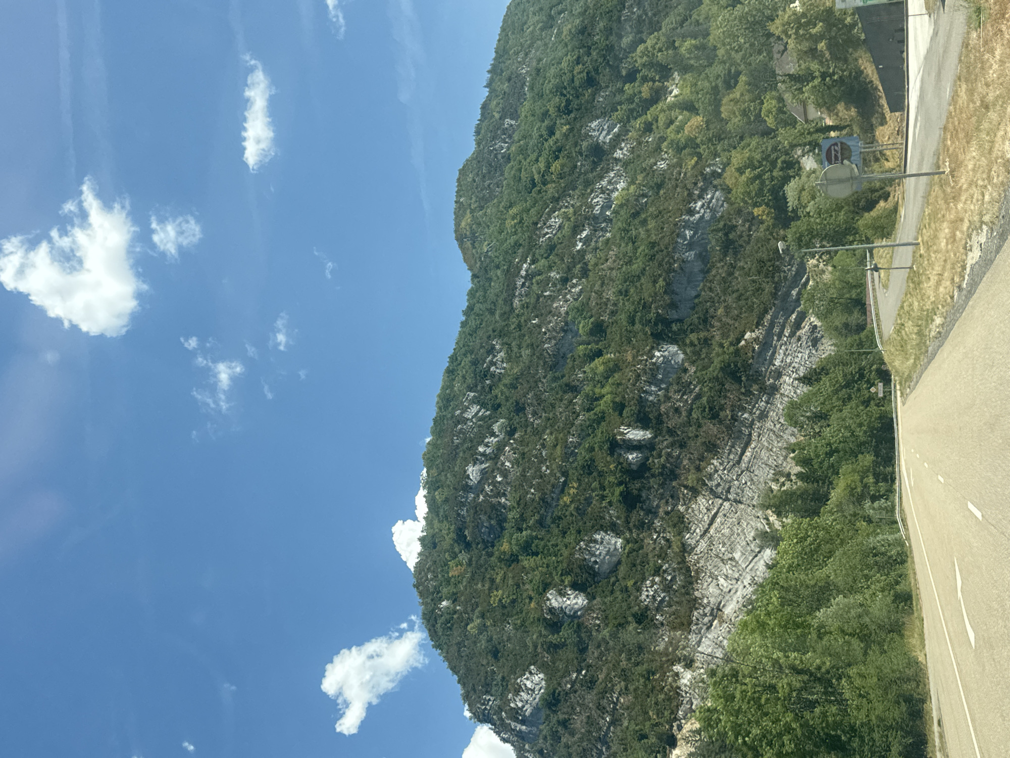 This image depicts a scenic view of a mountain range and a road in the vicinity of Villard-Saint-Sauveur, France. The mountain is covered in lush green vegetation, with exposed rocky strata visible on its slopes. Several white clouds dot the clear blue sky. A paved road with white lane markings curves through the foreground, suggesting a driving perspective. On the right side of the road, there are streetlights, road signs, and some buildings nestled amongst the trees. The overall impression is one of a natural landscape encountered while traveling.