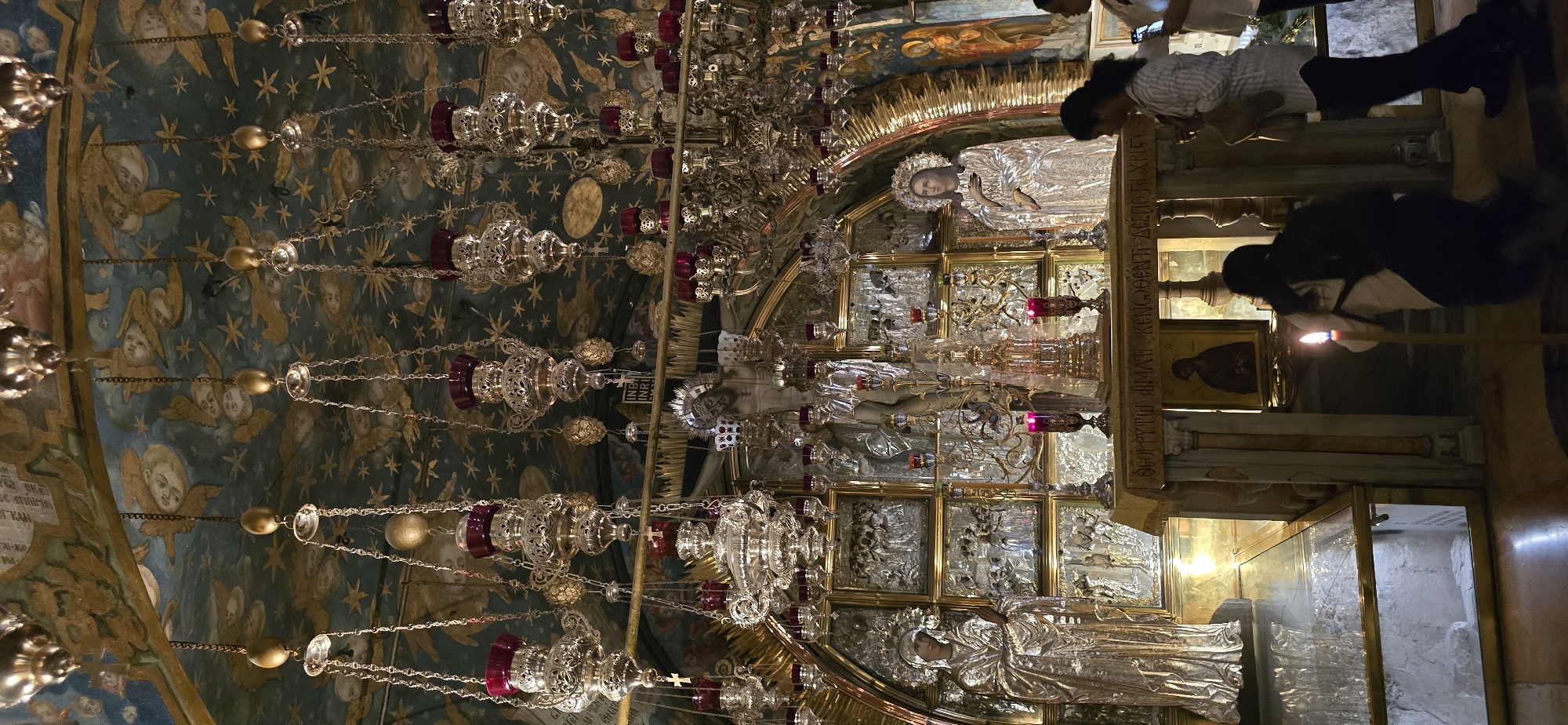The image depicts the interior of the Church of the Holy Sepulchre in Jerusalem, Israel. The scene is ornate and dimly lit, dominated by a large altar at the center. Above the altar, a gilded structure features a crucified Jesus figure, surrounded by numerous hanging oil lamps and ornate chandeliers. To the left of the altar is a silver statue of a robed figure, likely Mary, and to the right is another silver statue of a robed figure with a halo, possibly representing a saint or another religious figure. The walls and ceiling are richly decorated with frescoes of celestial motifs, including stars and angelic figures. In the foreground, two individuals are visible. One person, wearing a light-colored shirt and dark pants, is standing and looking towards the altar, holding a smartphone. Another person, wearing a light-colored top and dark pants, is kneeling in front of the altar, with a lit candle placed on the floor nearby. The altar itself has a stone surface with inscribed text and a framed religious icon. A glass-covered area in the lower left shows stone fragments, possibly representing a historical site within the church. The overall atmosphere is one of reverence and religious significance.