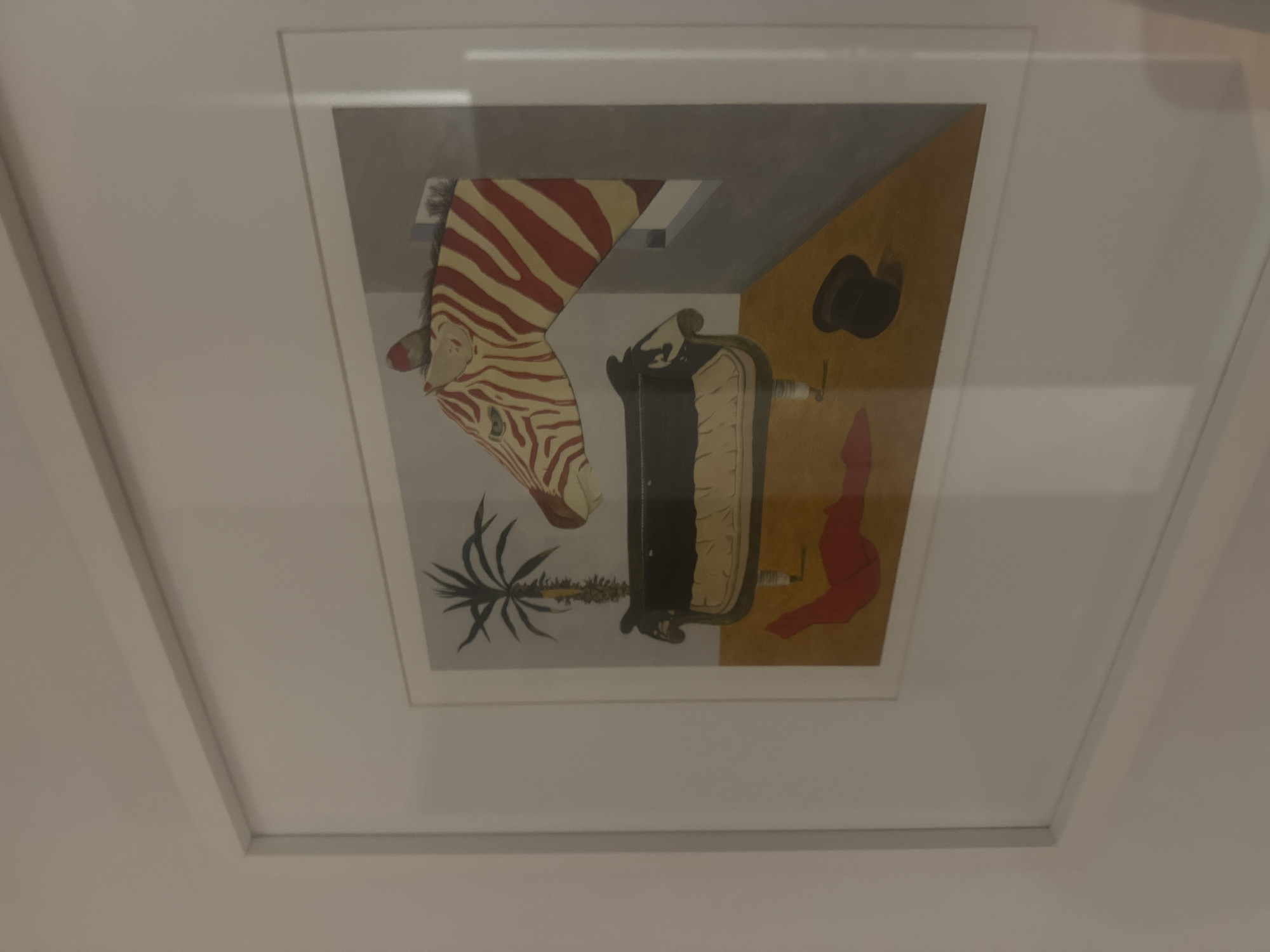 The image shows a framed artwork, likely a print, hanging on a white wall. The artwork features a surreal composition: a zebra head extending into a room-like space, with a portion of the head disappearing into a ceiling. The interior includes a couch, a palm tree, a hat, and a red, draped fabric. The colors of the interior are primarily gray and tan. It is located in Puebla, Mexico.