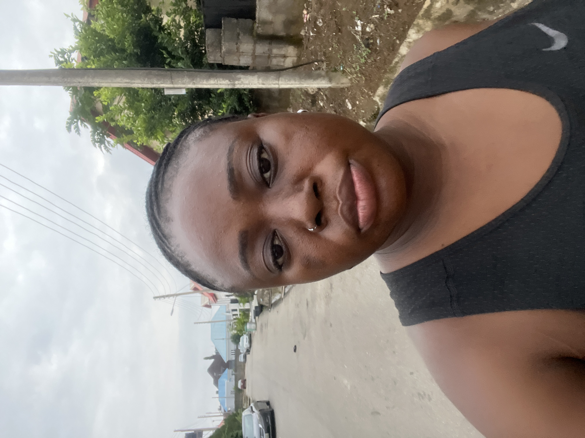 The image is a selfie taken outdoors on an overcast day in Abuja, Nigeria. A woman with dark skin and braided hair is in the foreground, facing the camera. She is wearing a black tank top. She has a nose ring and an earring. Her expression is neutral. In the background, a paved street stretches into the distance, lined with houses and trees. Several cars are parked along the street. A utility pole with overhead wires stands to the right of the woman. The overall environment appears to be a residential area.
