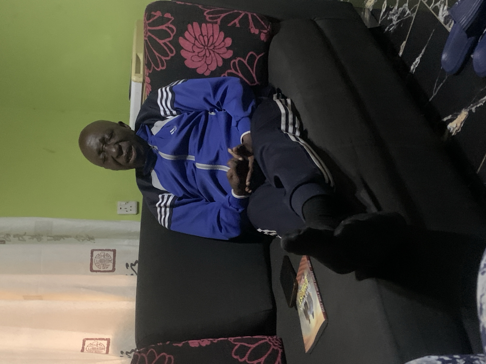 A bald, dark-skinned man is sitting on a black sofa in a room with a lime green wall and white curtains. He is wearing a blue tracksuit with white stripes. His eyes are closed and his mouth is open as if he is speaking or singing. His legs are crossed and his feet are bare. A book titled "Daily Walk with Christ 2020" and a black cell phone are on the sofa next to him. The floor is black marble with white veins. The scene is indoors, suggesting it could be a home or a hotel room in Jos, Nigeria.