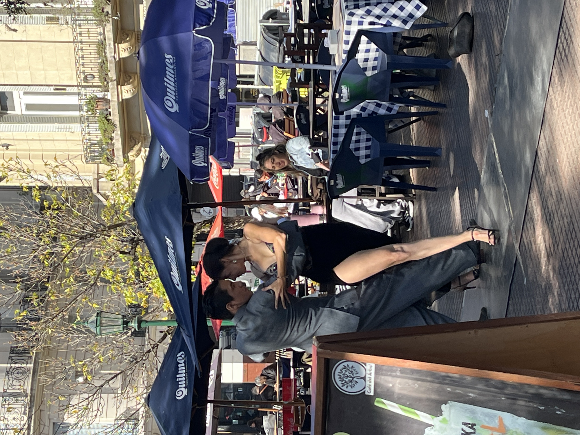 A medium outdoor shot depicts a tango performance occurring on a Buenos Aires, Argentina city street. A couple is actively engaged in a tango dance. The male dancer, dressed in a gray suit, is holding the female dancer, who is wearing a black dress. The dance is taking place near outdoor café seating. The background features rows of blue Quilmes-branded umbrellas and tables with checkered cloths. In the blurred background, pedestrians are visible. A cafe menu sign, featuring a tree logo and the words "Café del Arbol," is visible on the right side of the frame. The architecture in the background consists of multistory buildings with ornate balconies. A wrought-iron lamppost and a sparse tree are also present.