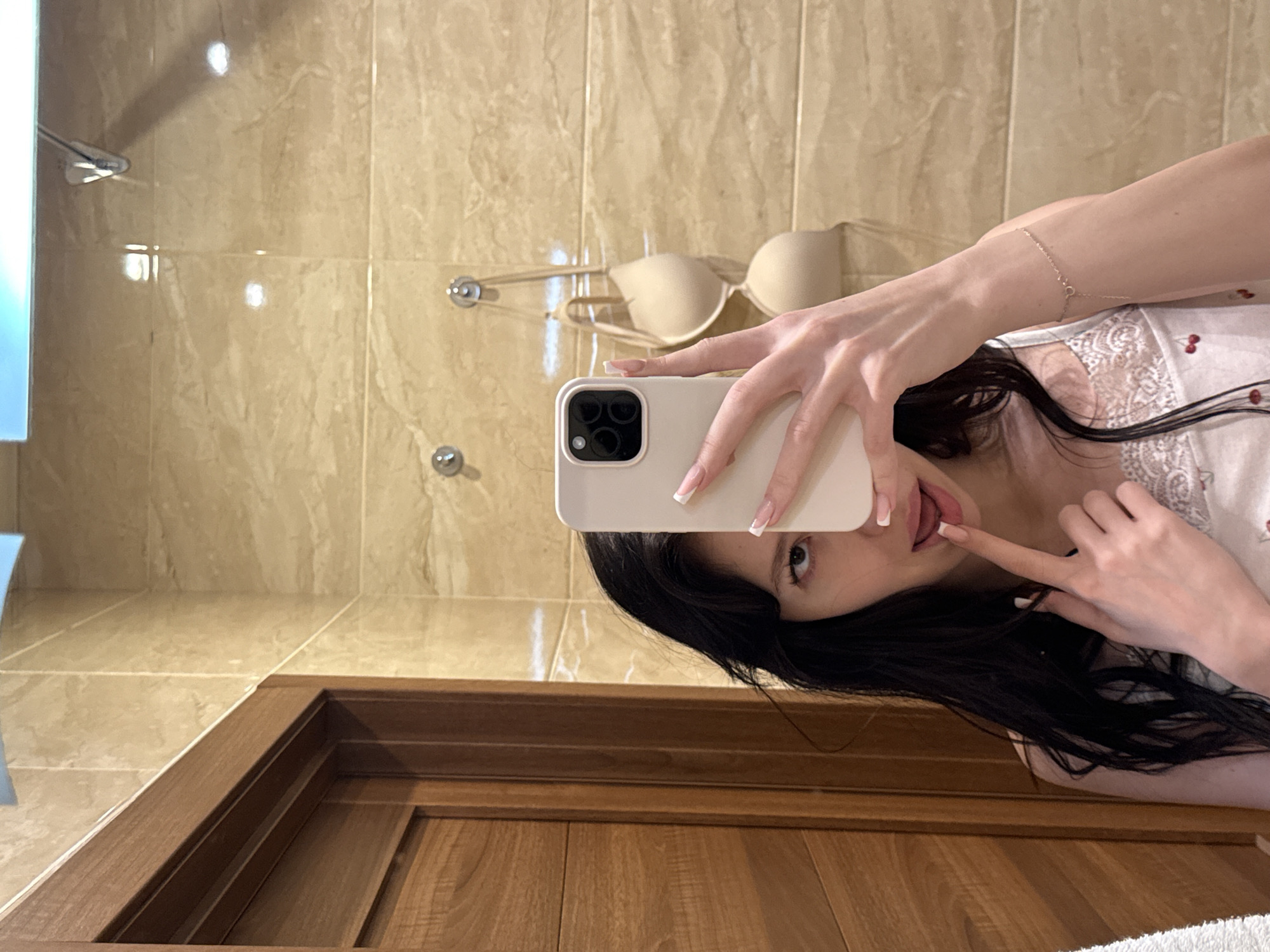 A young woman with long, dark hair and light-colored skin is taking a mirror selfie in what appears to be a bathroom in Khimki, Russia. She is wearing a white tank top with a cherry pattern and lace trim. Her left hand is holding up a white smartphone to take the photo, and her index finger is touching her lips. Her right hand is resting on her chest. The background shows a wall with beige marble tiles, a wooden door on the left, and two beige bras hanging on hooks on the wall. The lighting suggests an indoor setting, possibly a hotel bathroom.