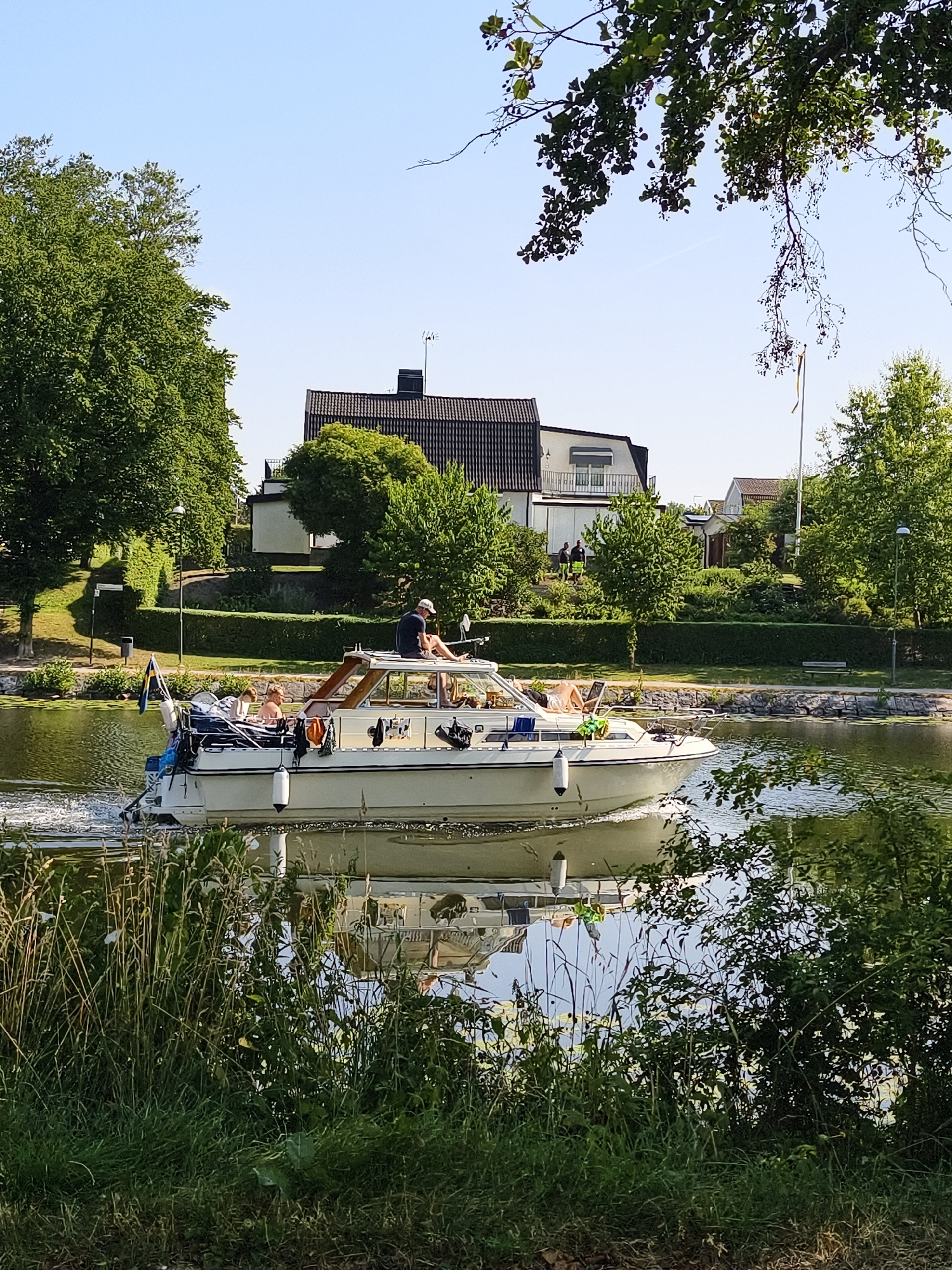 A white motorboat with a wooden cabin is cruising on a calm body of water, likely a canal or river. The boat has several people on board. A man in a dark t-shirt and white cap is sitting at the stern, facing away from the camera. Further forward, there are at least two other individuals on the deck, one of whom appears to be sunbathing. The boat is leaving a wake behind it, indicating it is in motion.
The surroundings suggest a suburban or semi-rural setting. On the left bank of the water, a lush green lawn slopes upwards towards a line of mature trees and a hedge. Beyond the trees, a modern-looking house with a dark tiled roof and white facade is visible. Another house, smaller and set back further, is also present. The right bank features dense greenery, including trees and bushes, with a stone wall at the water's edge. There are also indications of a pathway or promenade along the banks, with a bench visible on the right side. The sky is clear and blue, suggesting a sunny day. A Swedish flag is flying from the stern of the boat. The reflection of the boat and the surrounding greenery is clearly visible on the water's surface.
Given the presence of the Swedish flag and the architectural style of the houses, it is plausible this scene is located in Linköping, Sweden, possibly along the Kinda Canal or a similar waterway within or near the city.