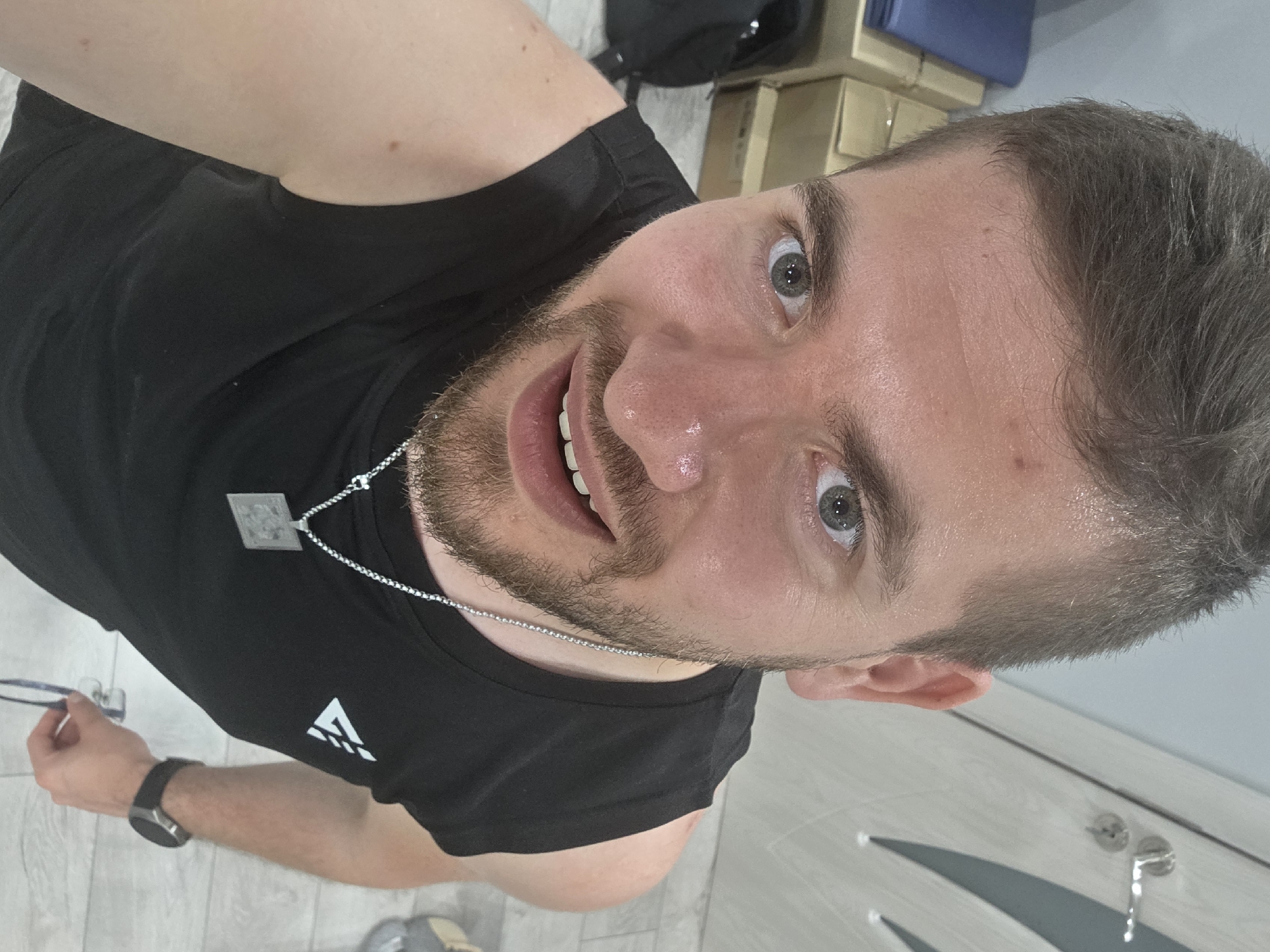 An overhead selfie captures a man with light skin, short dark blonde/light brown hair, and a trimmed beard, looking directly at the camera with wide, light blue-grey eyes and a pleasant, open-mouthed smile. His face shows a slight sheen, possibly from sweat.
He is dressed in a black sleeveless shirt with a white, stylized logo on the left chest, resembling an upward-pointing arrow or mountain peak. A silver chain necklace with a square, engraved pendant rests on his chest. On his left wrist, he wears a black digital watch, and he holds a pair of glasses in his left hand.
The indoor setting, in Yagodovo, Bulgaria, appears to be a casual space, possibly a changing room or a personal area. The background includes a dark bag, several light brown cardboard boxes, and a blue item. The floor is a light-colored tile or wood laminate. A reflective surface, likely a mirror, is visible in the lower right, showing a reflection of a grey door or wall with hardware.