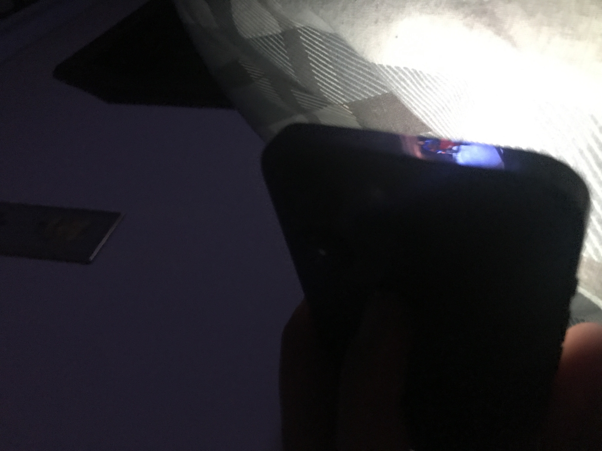A dark-colored smartphone is held in someone's hand, reflecting light on its top edge. The background includes a dark surface and a gray and white patterned surface, possibly a bedspread or fabric. A dark rectangular object is also visible in the background. The location and city of Yerevan, Armenia, cannot be determined from this image.