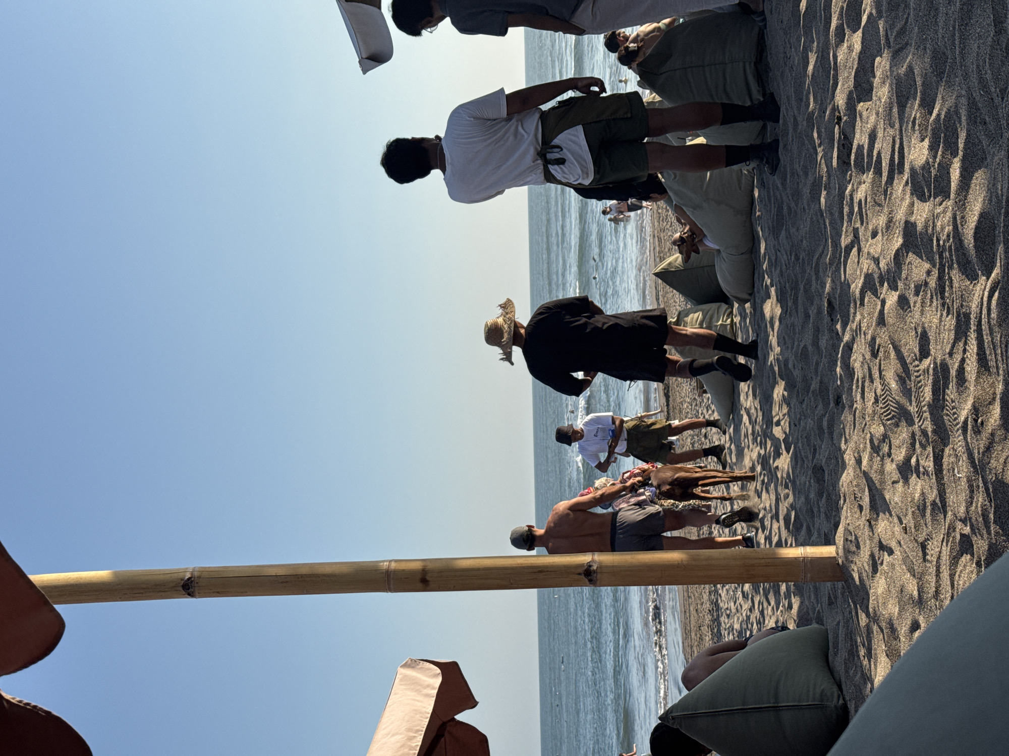 This image depicts a beach scene in North Kuta, Indonesia, likely during daylight hours. The foreground is dominated by sandy beach, with several large, olive-green beanbag chairs scattered around. A tall, bamboo pole stands prominently on the left, supporting an unseen structure that casts a shadow.
In the middle ground, a group of people are visible on the beach. A man with his back to the camera, wearing grey shorts and a dark cap, is walking a brown dog on a leash. To his right, another man in a white t-shirt and olive-green shorts is interacting with something held in his hand, possibly a phone or a small item. Further to the right, a man in a black shirt and a straw hat is standing, facing the ocean. In the background, the ocean stretches out, with gentle waves and a clear, pale blue sky above. Several other individuals are scattered along the shoreline and in the shallow water, some appearing to be swimming or wading.
The overall impression is a relaxed, beachside atmosphere with people enjoying the coastal environment. The lighting suggests it could be late afternoon or early morning, given the warm tones and the shadows.