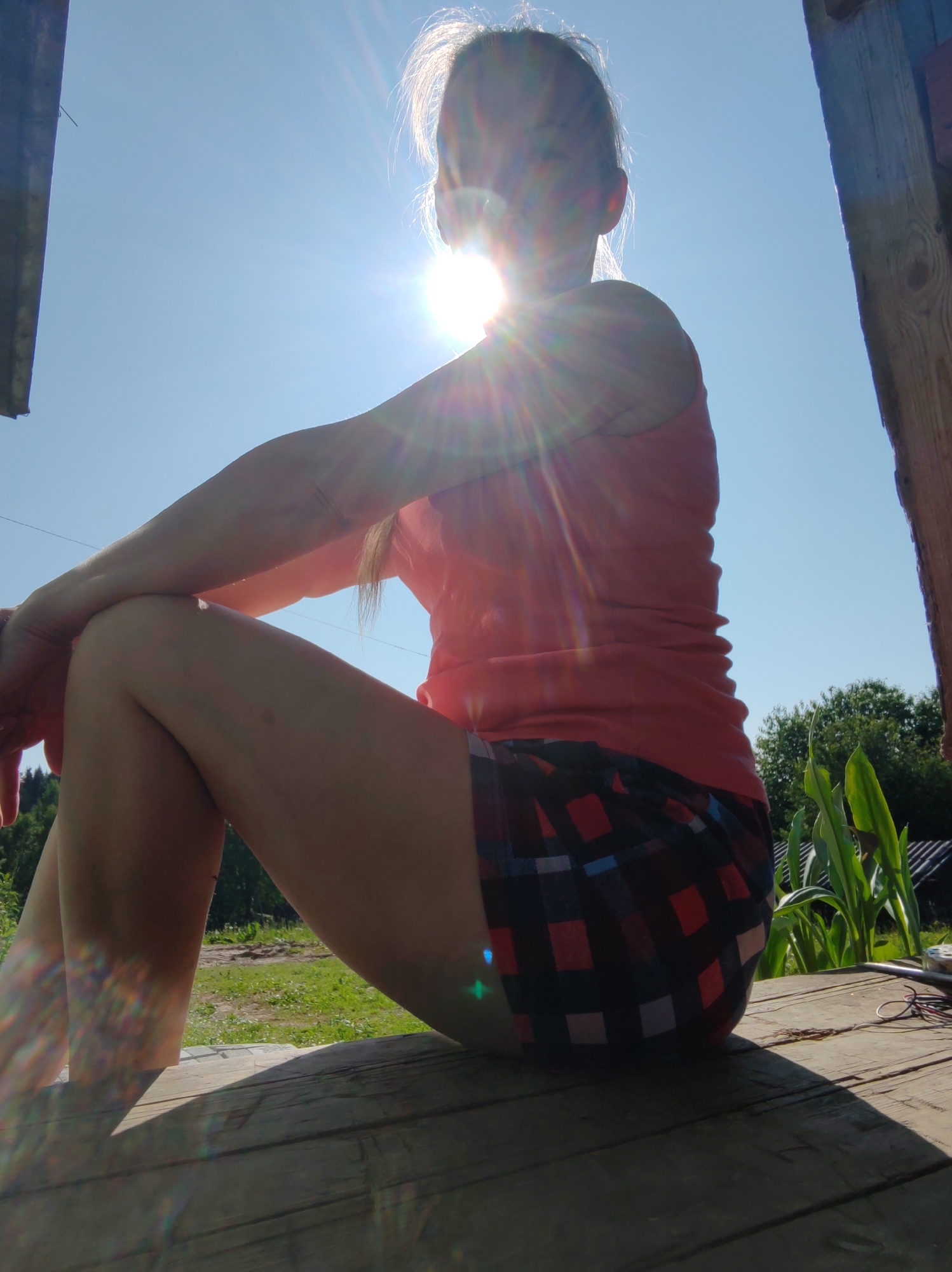 A person is sitting on a wooden platform outside, with the sun shining brightly in the sky, creating lens flares. The person is wearing a coral-colored tank top and plaid shorts. Their arms are crossed, and their legs are bent. In the background, there is green grass, trees, and a roof structure. The overall scene suggests a casual outdoor setting, possibly in a rural or suburban area of Чухлэм, Russia.