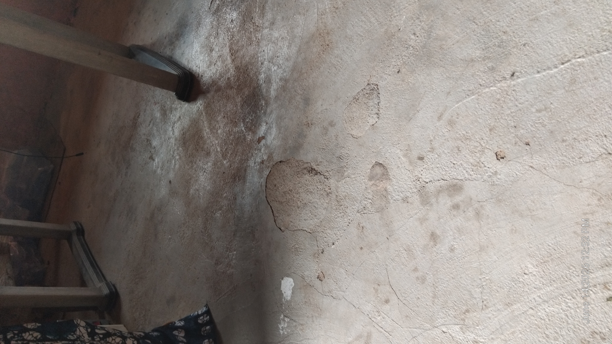 A low-angle, eye-level shot captures a portion of a concrete floor, likely in an indoor setting within Howayi, Nigeria. The floor exhibits signs of wear and damage, with several areas of chipped or missing concrete, particularly a large, irregular patch in the center and smaller ones scattered around. Cracks are also visible across the surface. In the upper left corner, a dark fabric with a white, abstract pattern is partially visible, suggesting the presence of clothing or upholstery. Two wooden table legs, one on the left and one on the right, are present, indicating the presence of furniture. The lighting appears dim and natural, casting subtle shadows. A timestamp at the bottom left corner reads "June 14, 2026 12:52 PM".
