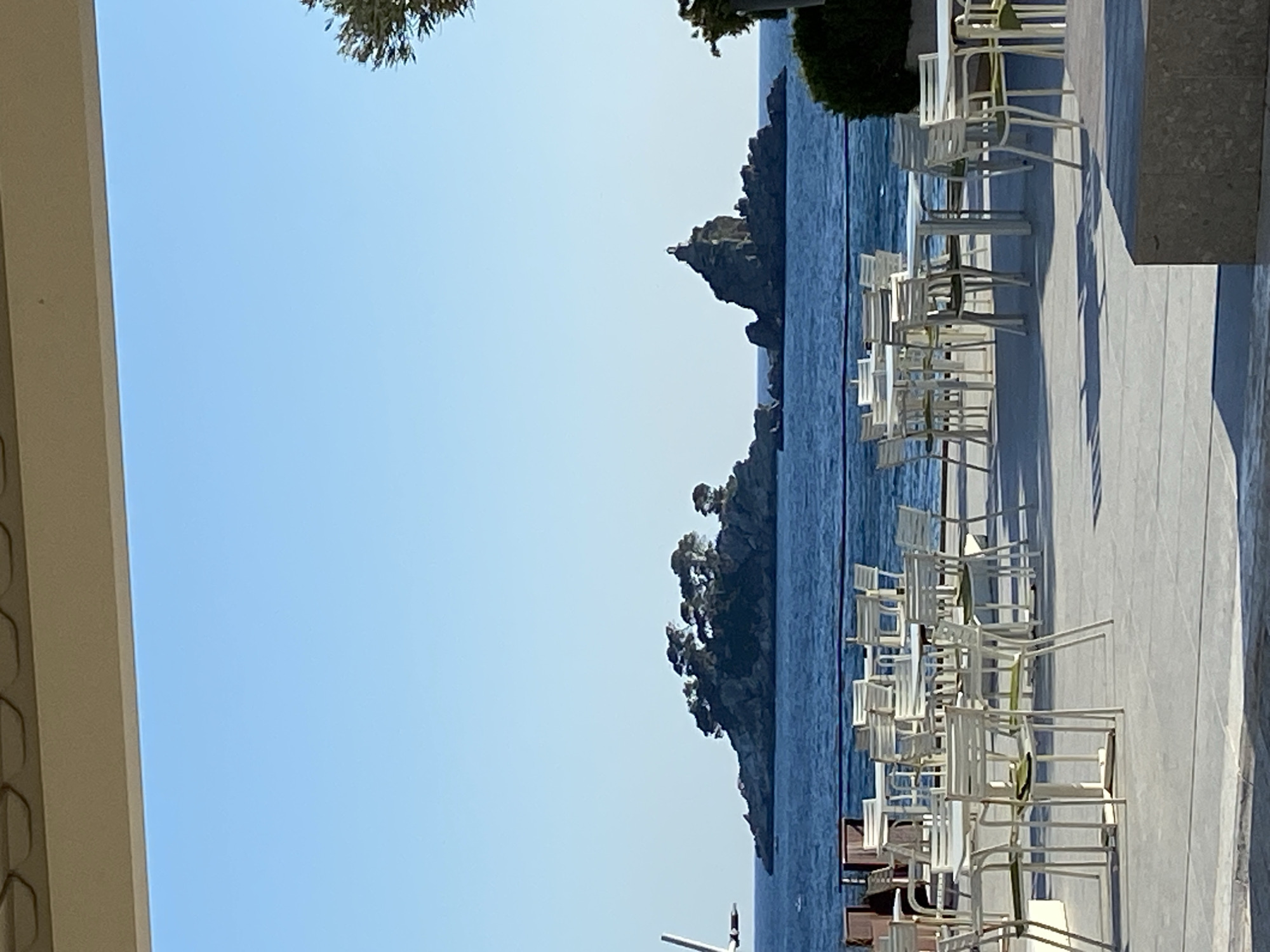 The image shows a seaside view in Petrovac na Moru, Montenegro. In the background, there are two small rocky islands in the sea. In the foreground, there is a patio with white chairs and tables. The sky is clear blue. The image is taken from a slightly elevated position under a building.