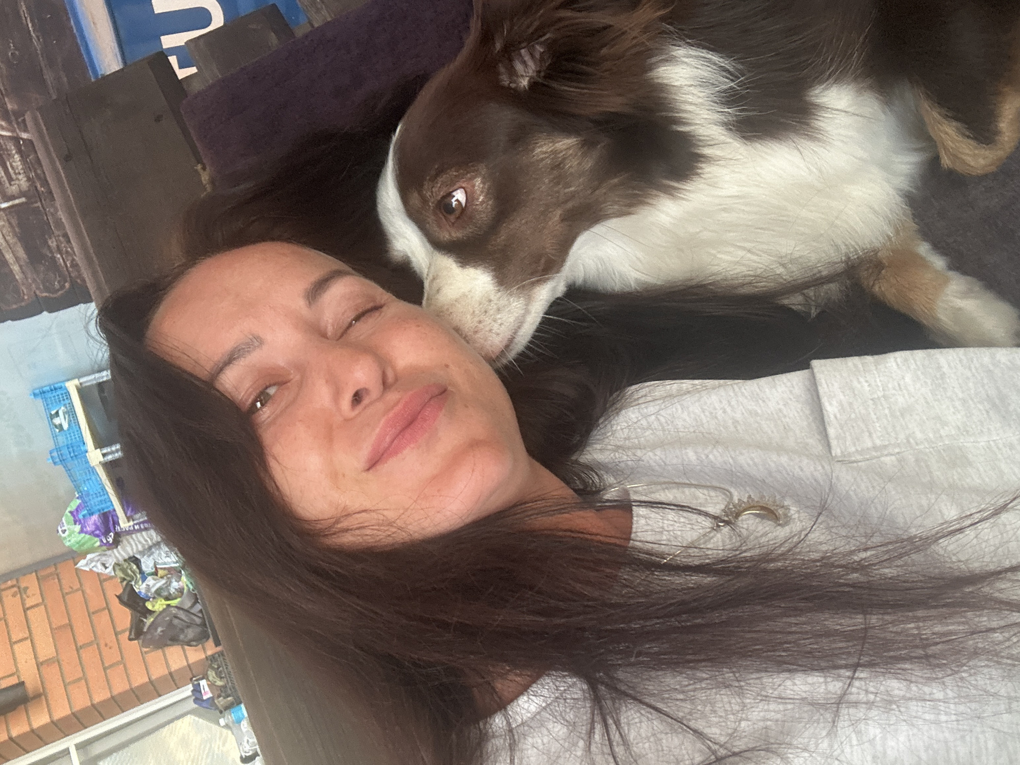 The media file shows a close-up, overhead shot of a woman and a dog. The woman is lying down, with her eyes closed and a slight smile on her face, appearing relaxed and content. Her long, dark hair is spread out around her. A brown and white dog is nuzzling her cheek, with its head resting near her face. The dog's eyes are open and it appears to be looking at the camera. They are both on a dark surface, possibly a couch or bed, with a light-colored garment worn by the woman visible. In the background, there are some shelves with various items, including blue crates and plastic bags, suggesting an indoor setting with some clutter. Brickwork and a window frame are visible in the lower left portion of the image. The overall impression is one of affection and closeness between the woman and her pet. The lighting suggests it could be daytime, possibly indoors with natural light. There is no visible text in the image.