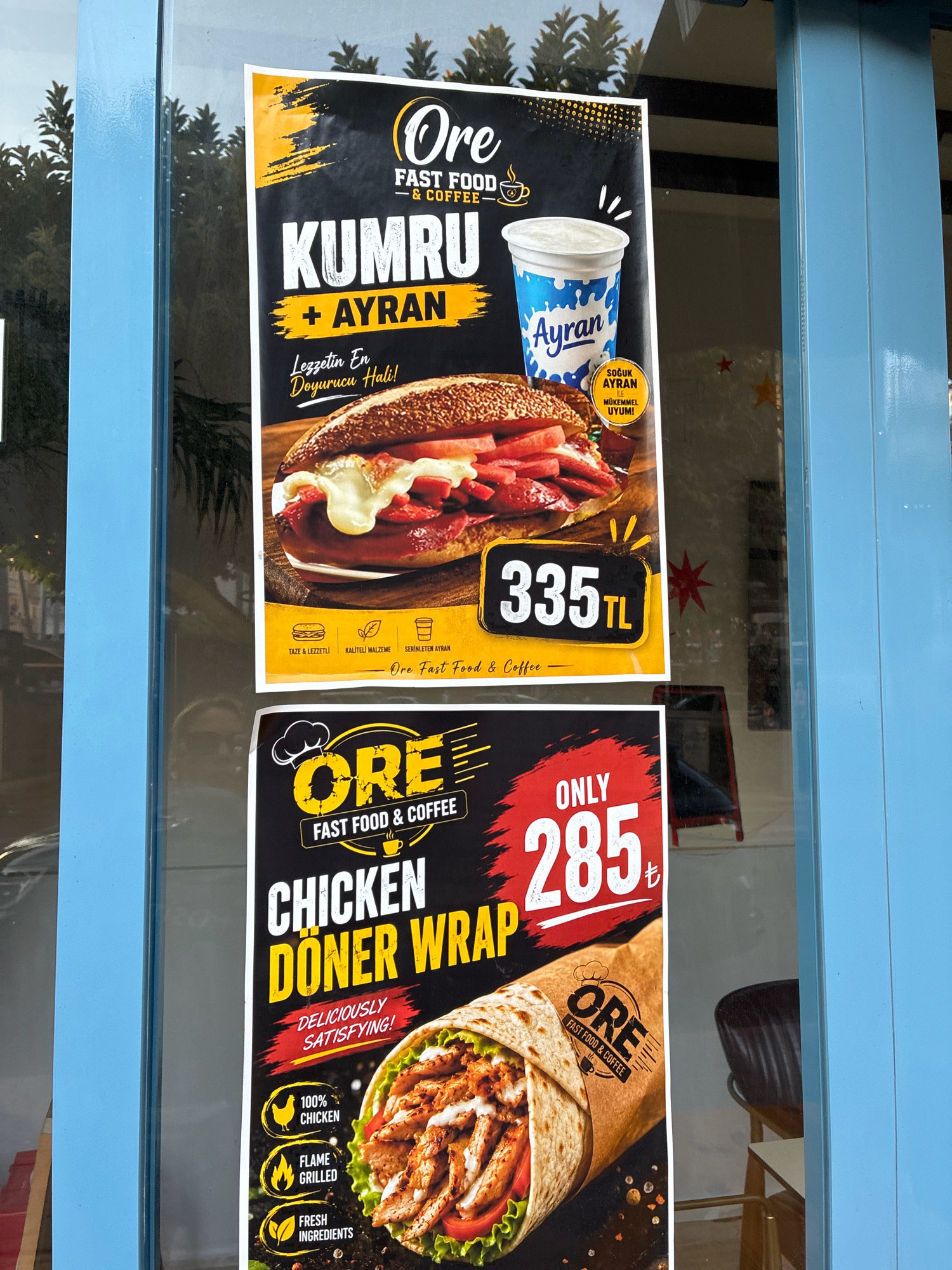 The image shows two advertisements for Ore Fast Food & Coffee displayed on a glass storefront. The storefront is painted a light blue and is likely located in Alanya, Turkey, based on the currency symbol "TL" (Turkish Lira) used in the advertisements and the context of common storefronts in tourist areas of Turkey.
The top advertisement features a "Kumru + Ayran" meal deal for 335 TL. The advertisement includes an image of a sandwich filled with sausage and cheese, a cup of ayran (a yogurt drink), and text in Turkish describing the food and drink. Below this are icons representing a sandwich, ingredients, and a coffee cup, with accompanying text in Turkish.
The bottom advertisement, partially obscured by the top one, advertises a "Chicken Doner Wrap" for 285 TL. This advertisement features a close-up image of a doner wrap, with icons indicating "100% Chicken," "Flame Grilled," and "Fresh Ingredients." The Ore Fast Food & Coffee logo is also visible on this advertisement.
The advertisements are hung on the inside of the glass, with the exterior environment reflected on the glass surface. Trees and foliage are visible in the reflection, suggesting an outdoor setting. No people are clearly visible in the frame. The lighting suggests it is daytime, with sunlight illuminating the scene.