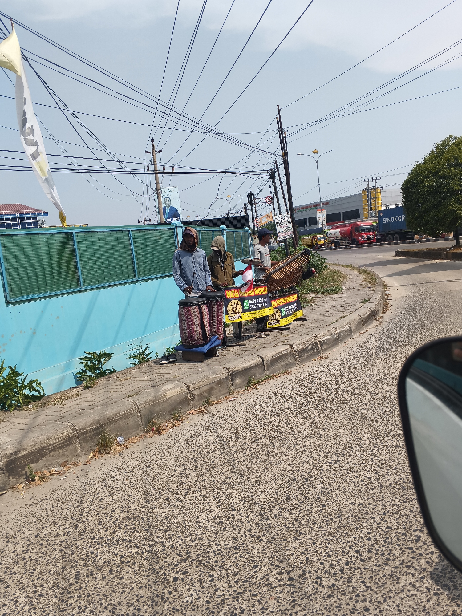 This is a street-level photograph taken in Bandar Lampung, Indonesia, on a sunny day. In the foreground, a paved road is visible, with the reflection of a car's side mirror on the right. On the left side of the road, there is a sidewalk bordered by a low concrete curb. A bright blue wall runs along the back of the sidewalk, with a green slatted fence on top.
Three individuals are present on the sidewalk. The person on the left, wearing a blue shirt and a headscarf, stands beside two large cylindrical drums. The drums are adorned with colorful, patterned fabric. To their right, a person wearing a brown jacket and a hooded garment is also present. Further to the right, a third individual in a light-colored t-shirt is seen playing a wooden musical instrument, possibly a gamelan or a similar percussive instrument, which is placed on a stand. Two black and yellow signs are positioned near the musical instrument, displaying text and contact information.
In the background, a white flag with yellow accents is flying from a pole. Overhead, a complex network of electrical wires crisscrosses the sky. A poster of a man in a suit is visible, along with several commercial buildings and vehicles, including trucks. The overall scene depicts a street environment with a temporary setup for musical performance on the sidewalk.