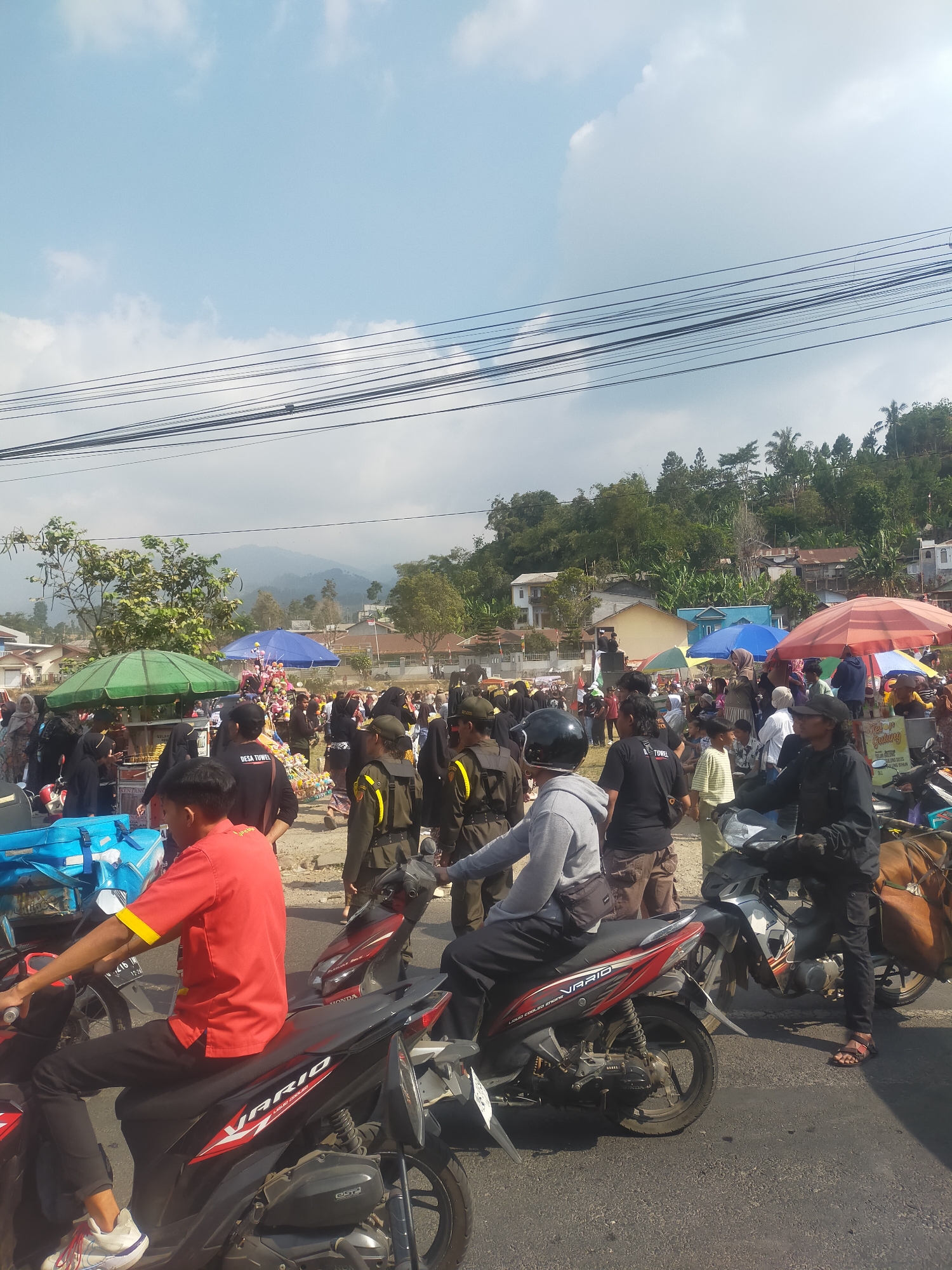 The image captures a bustling street scene in the city of Tuwel, Indonesia. The foreground is dominated by several parked motorcycles, with one occupied by a person in a red shirt and black pants. Further back, a dense crowd of people are gathered, many under colorful umbrellas, suggesting a market or event. Several individuals are wearing what appear to be official uniforms, possibly security personnel. Behind the crowd, the landscape rises into lush green hills dotted with houses, indicating a semi-urban or rural setting. The sky is a clear blue with scattered white clouds. Power lines are visible crisscrossing the upper portion of the image.