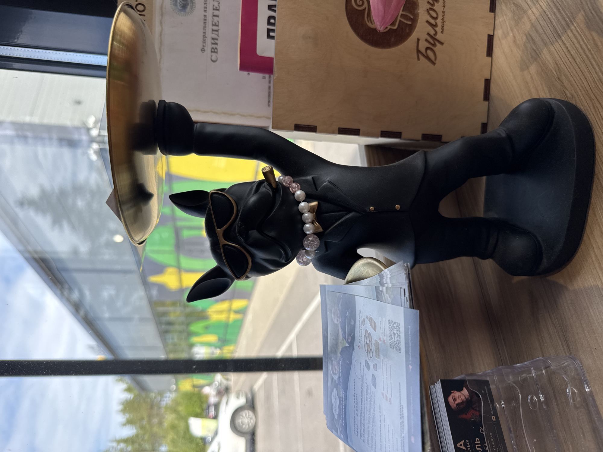A black statue of a French bulldog wearing sunglasses, a pearl necklace, and a suit, holding a golden tray, stands on a wooden shelf next to a window. Outside the window, trees, a white car, and a building with a colorful mural are visible. On the shelf, there are brochures, a business card holder with a business card, and a wooden box with Russian lettering. The scene appears to be indoors, possibly in a cafe or shop, in the city of Finskiy Briz, Russia.