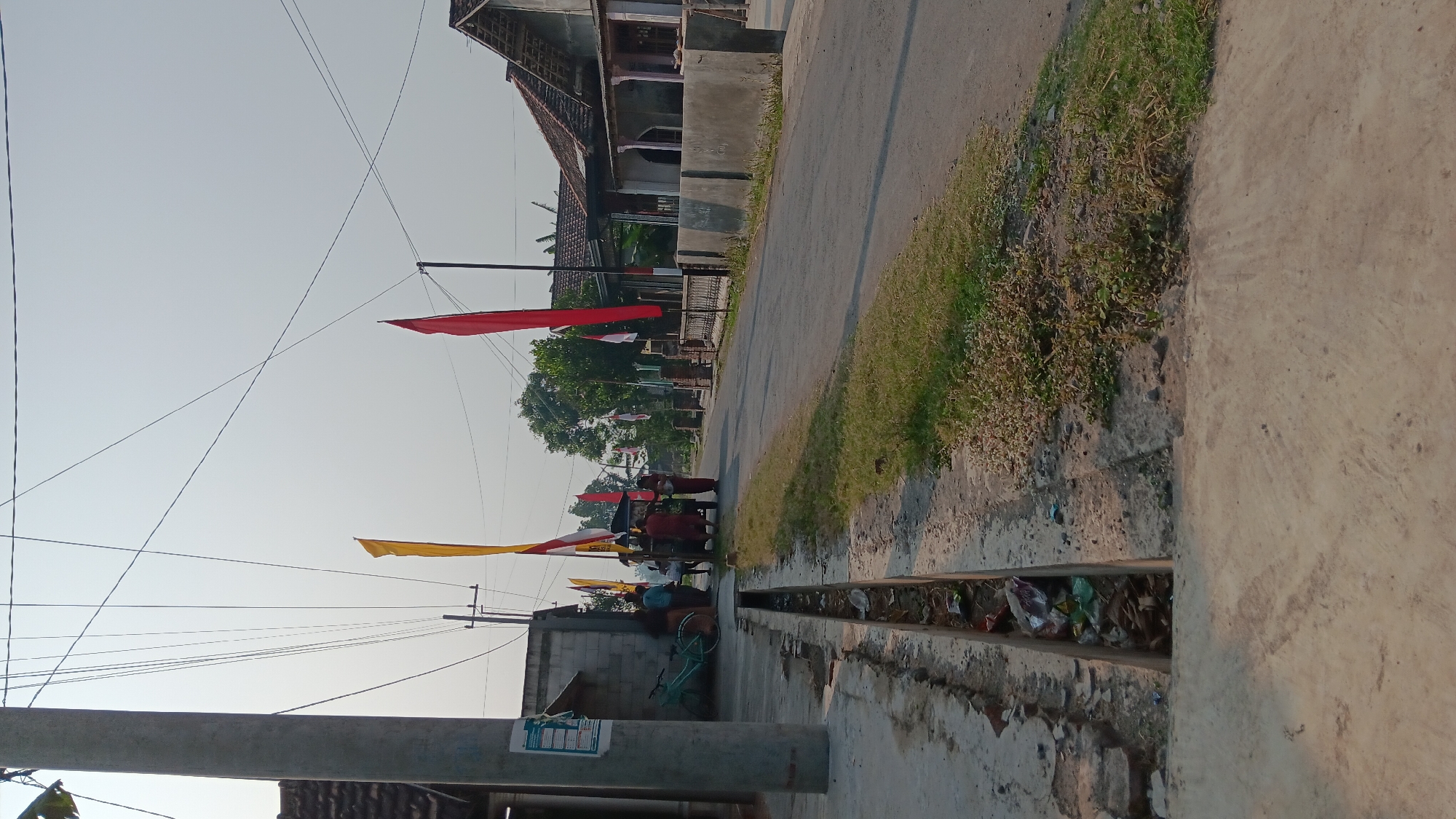 This image depicts a street scene in Sonobekel, Indonesia, likely on a clear day with the sun casting shadows. A paved road runs diagonally through the frame, bordered by a grassy verge and a concrete sidewalk. A narrow, open drainage ditch runs alongside the sidewalk, containing visible debris and trash. On the left side of the road, a gray concrete utility pole stands tall, adorned with multiple electrical wires crisscrossing overhead. Adjacent to the pole is a low, rough-hewn concrete building. A light blue bicycle is leaned against the building. Several people are gathered near a small, multi-passenger vehicle, possibly a bemo or similar local transport. Red and yellow flags are displayed along the road, some on poles and some attached to the vehicle, suggesting a local celebration or event. Houses with tiled roofs are visible on the right side of the road, separated from the street by low concrete walls. The overall impression is of a quiet, rural-to-suburban street in Indonesia.