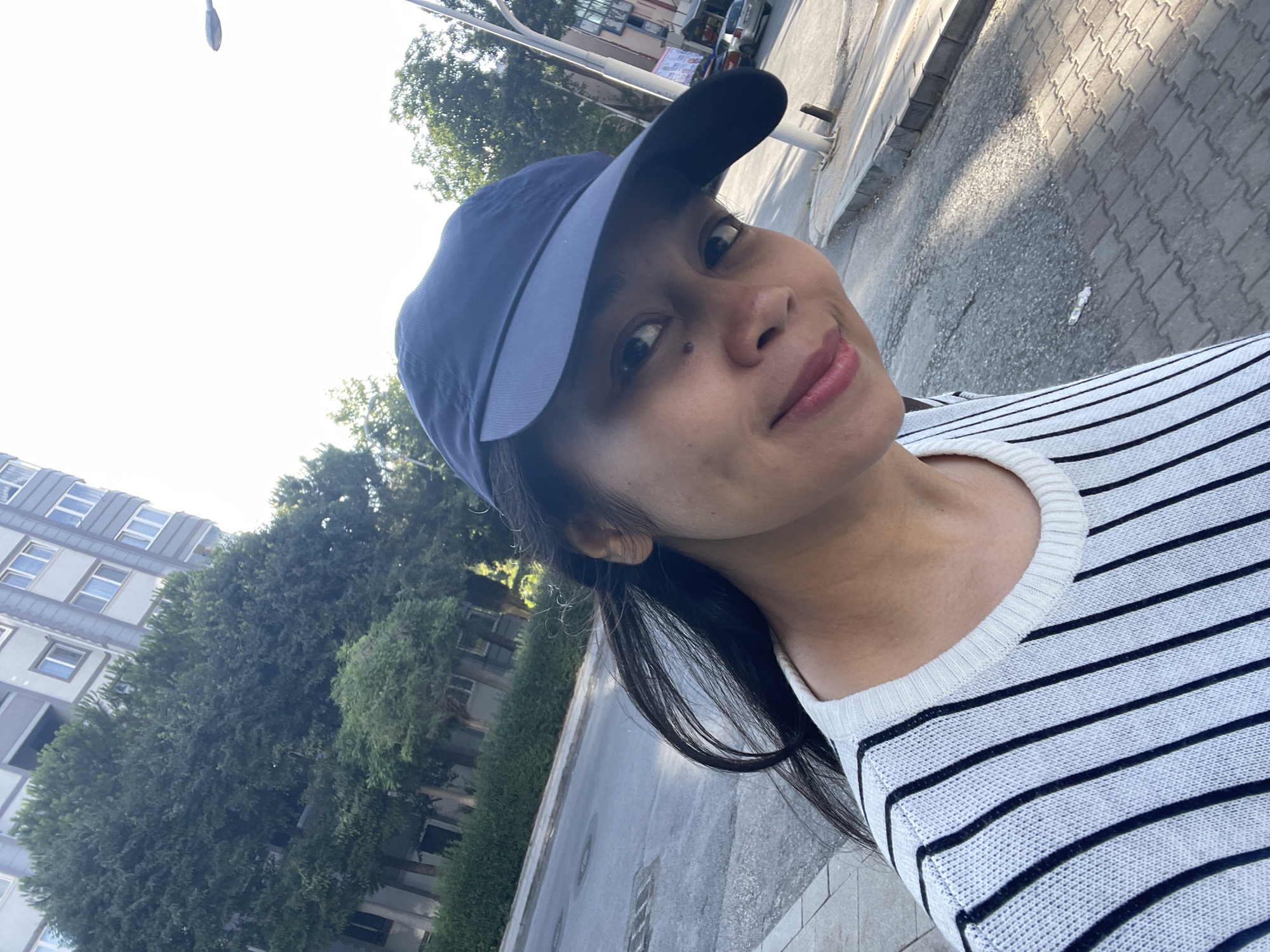 A woman wearing a blue baseball cap and a white and black striped sweater is posing for a selfie outdoors. She has dark hair and is smiling at the camera. The background shows a street, a sidewalk, trees, and a building. The scene appears to be in the city of Başakşehir, Turkey, based on contextual information not visible in the image itself. The lighting suggests it is daytime, possibly late afternoon, with the sun casting shadows on the ground.