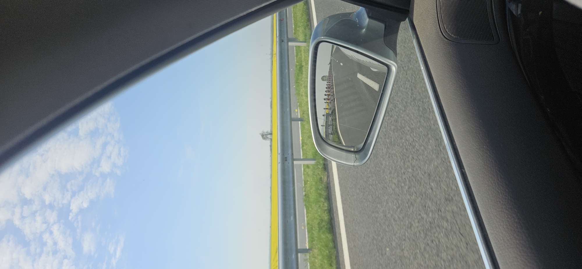 The image, taken from inside a car looking out the passenger window, depicts a bright, sunny day on a highway. The sky, a clear blue with scattered white cumulus clouds, occupies the upper-left portion of the frame (when viewed with the road horizontally).
The scene outside the car features a multi-lane asphalt road, bordered by a metal guardrail. Beyond the guardrail, a vibrant field of yellow crops, characteristic of rapeseed or canola, stretches into the distance, with a single, small tree visible further back.
A significant detail is the car's exterior side mirror, positioned on the right side of the frame. Its reflection clearly shows the road receding behind the vehicle, lined by numerous small red and white posts or barriers. A small crack is noticeable on the silver casing of the side mirror. Parts of the car's interior, including the dark grey roof lining, window frame, and a section of the door panel with a speaker grille, are visible in the foreground.
The bright lighting suggests it is daytime, likely morning or early afternoon, with clear weather. The expansive yellow fields point to a rural or agricultural setting, consistent with the provided location context of Загорци, Bulgaria. No visible text is present in the image.
