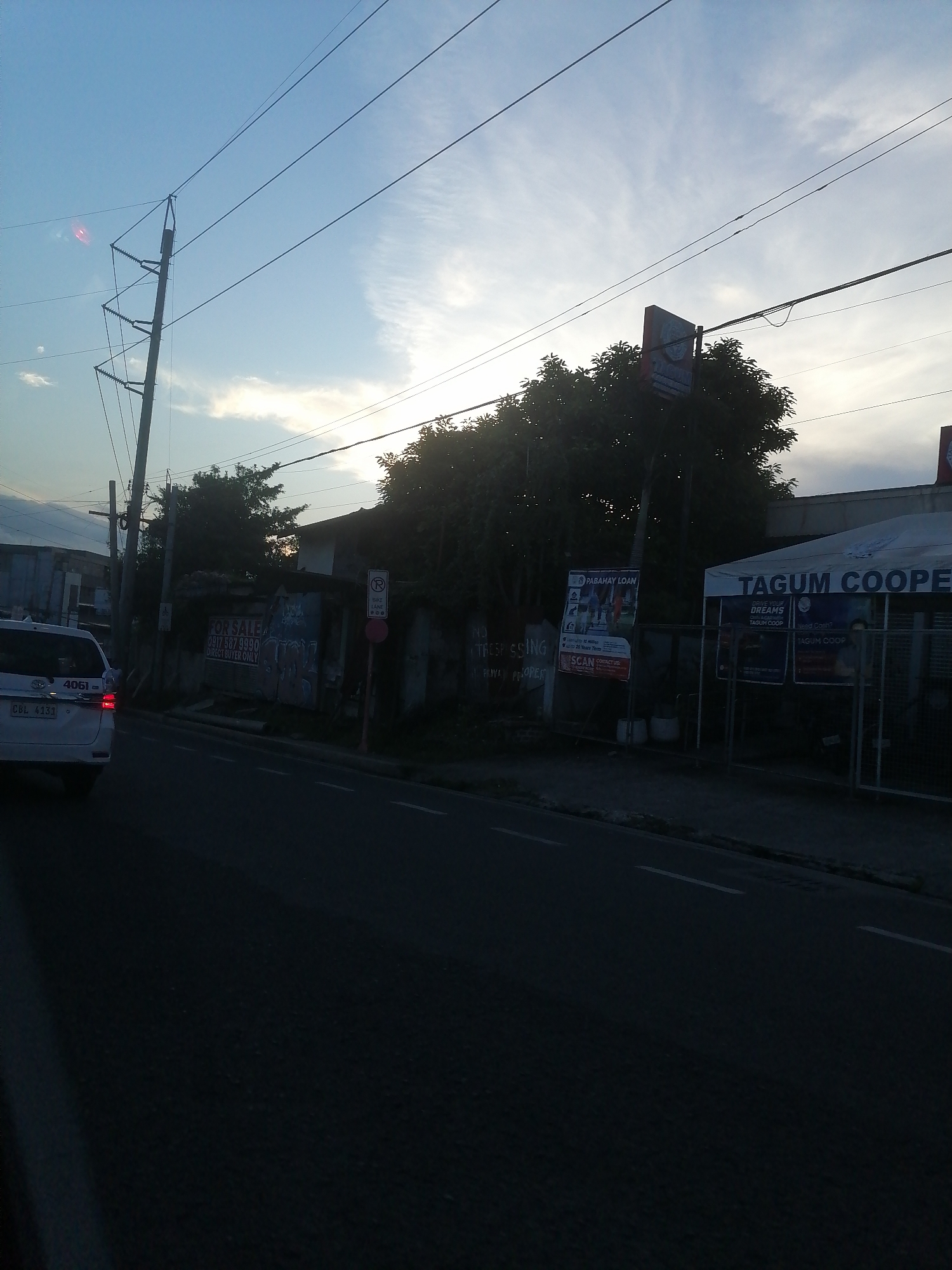 This image depicts an urban streetscape at what appears to be either dawn or dusk, characterized by soft, ambient light illuminating the sky while foreground elements remain largely shadowed. The scene is likely located in Davao City, Philippines, indicated by local signage such as "TAGUM COOPE" (referring to Tagum Cooperative, which has branches in Davao City), "PABAHAY LOAN" (Tagalog for Housing Loan), and a Philippine mobile number prefix (0917) on a "FOR SALE" sign.
The sky occupies the upper half of the frame, displaying a gradient of light blue transitioning to bright white near the horizon, with wispy white clouds scattered across. Multiple overhead power lines and communication cables traverse the sky, supported by a prominent utility pole on the left side of the road.
In the lower left, the rear section of a white vehicle, possibly a taxi or private car, is partially visible, displaying the unit number "4061" and license plate "CBL 4131." Adjacent to the road, on the left, is a wall or fence with graffiti, behind which stands a structure bearing a "FOR SALE" sign. The sign includes a contact number "0917 587 9990" and the text "DIRECT BUYER ONLY." Further down the road, a red post with a circular regulatory sign for "BIKE LANE / NO STOPPING / NO PARKING" is visible. Dense foliage and trees line the left side of the street, partially obscuring buildings.
On the right side of the road, a white tent-like structure or facade prominently features the text "TAGUM COOPE," indicating a branch of Tagum Cooperative. Several promotional posters are affixed to the fence or wall in front of this structure, with one clearly advertising a "PABAHAY LOAN." More trees and dark foliage are visible behind the cooperative building.
The asphalt road in the foreground is dark due to shadows, with faint white dashed lane markings visible. No individuals are discernible in the frame. The overall impression is an active urban environment with commercial establishments and residences, typical of a Philippine city street.