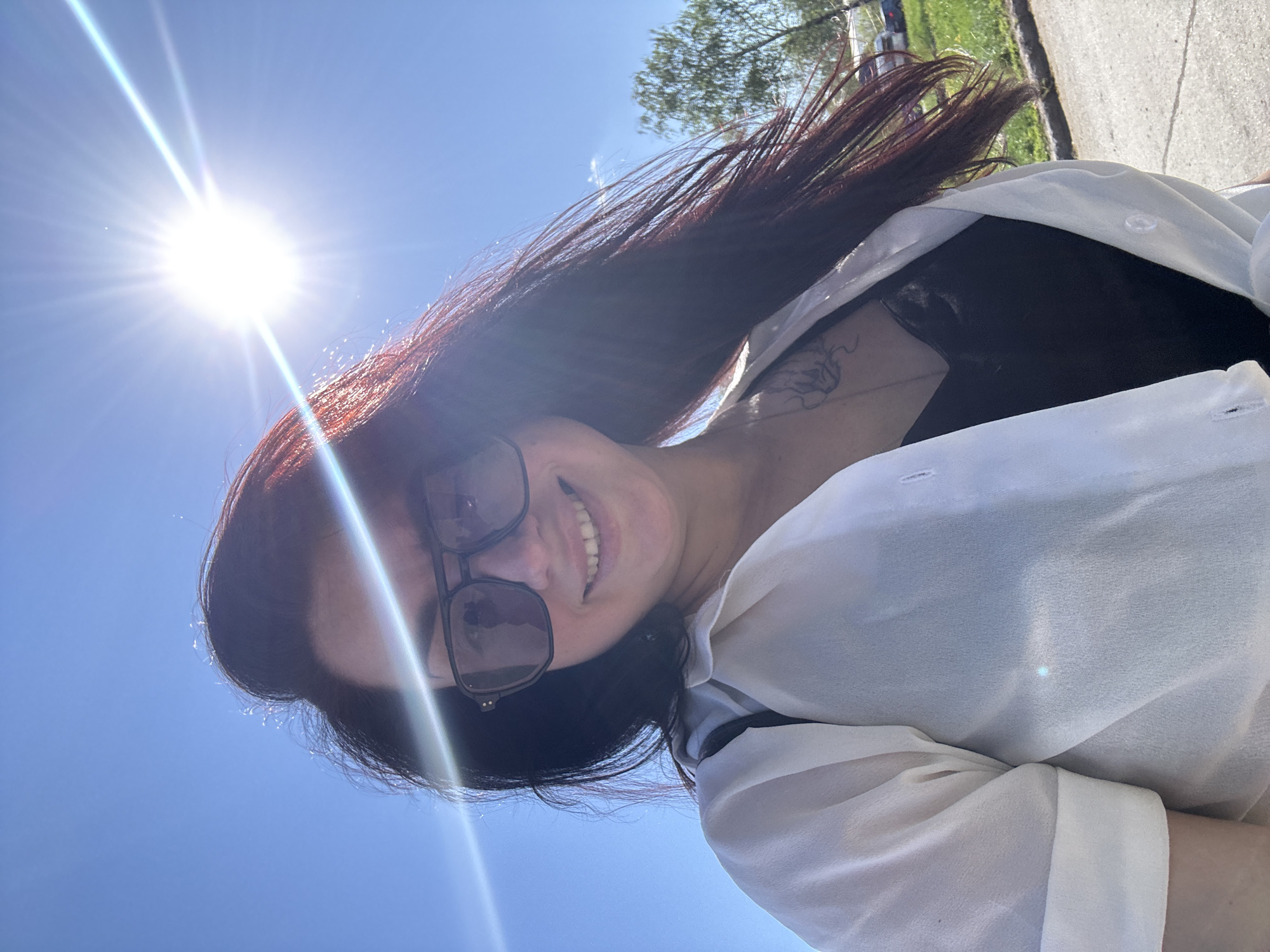 A woman stands outside on a sunny day. She wears sunglasses and a white blouse over a dark top. She is smiling. The background is a bright blue sky with the sun prominently visible. There is also a tree and a sidewalk. The location is Unknown, Unknown.