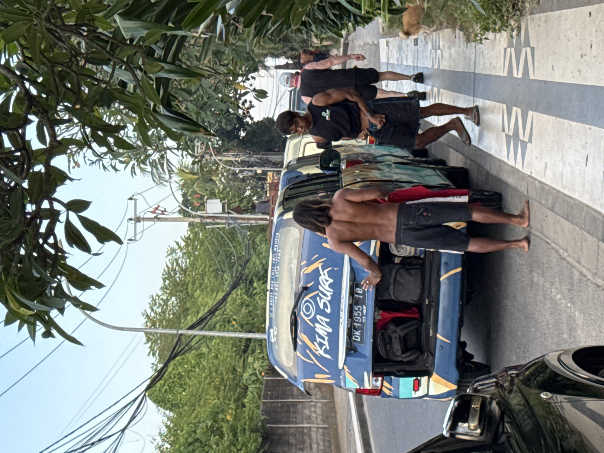 The scene depicts a street in Kuta, Indonesia, during daylight hours. In the foreground, a dark-colored vehicle is partially visible on the left. To its right, a blue van with a surf-themed livery, featuring "KIMA SURF" branding in white and orange/yellow graphics, is parked on the side of the road with its rear hatch open. The van's license plate reads "DK 1955 IO". A sticker on the rear window states: "IF I DRIVE BAD CALL THIS NUMBER +62 821-3154-7663". The open hatch reveals several pieces of luggage, including at least two dark backpacks and a red bag.
Two men are positioned near the rear of the blue van. The man on the left, with long dark hair, is shirtless and wears dark shorts, standing barefoot on the sidewalk with his left hand resting on the van's tailgate. A red garment is draped over his left arm. The man to his right, wearing a black tank top and dark shorts, is also barefoot or wearing minimal footwear, and appears to be adjusting his shorts or holding a small item in his hands.
Further down the sidewalk to the right, an older man wearing a black tank top, dark shorts, and a light-colored cap walks away from the viewer. Another man in dark attire is visible further in the background on the sidewalk. A small, light brown dog stands on the far right side of the sidewalk.
The environment includes a paved asphalt road, and a light-colored tiled sidewalk with a central dark grey stripe featuring a repeating geometric pattern. Abundant green foliage and trees line both sides of the street and extend into the background. Overhead, numerous black power lines crisscross the light blue sky, and a curved metallic streetlight pole is visible. A low concrete wall is present on the left side of the street, and a higher, textured wall is behind the blue van.