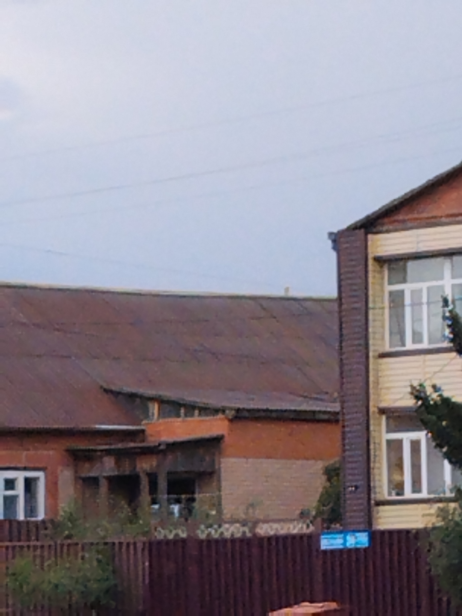 The image depicts an outdoor scene in Kokshetau, Kazakhstan, featuring two buildings and a wooden fence in the foreground. On the left, a long, low building with a rust-colored corrugated metal roof is visible. This building has a red brick facade and at least one white-framed window. To the right, a taller, two-story building with a yellow-paneled exterior and a reddish-brown roof is present. This building has multiple white-framed windows. A dark brown wooden fence runs horizontally across the bottom of the image, partially obscuring the lower portions of the buildings and some greenery. In the background, the sky is overcast with a pale, washed-out appearance. There are no people or animals visible in the frame. The overall impression is that of a residential or possibly institutional area, with elements suggesting a somewhat aged or rural setting.